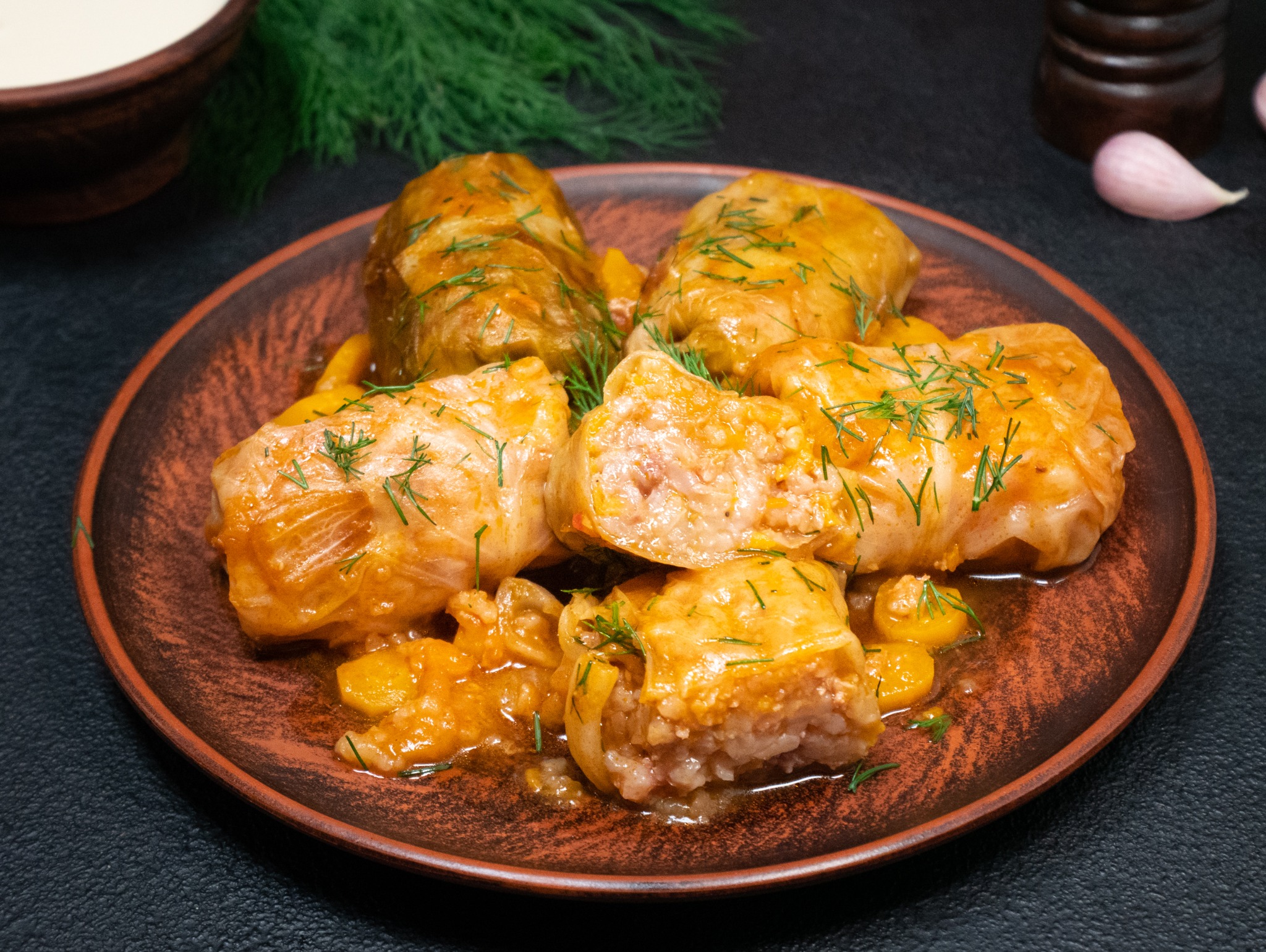 Tradycyjne gołąbki na brązowym talerzu, posypane świeżym koperkiem, z sosem i dodatkami. W tle przyprawy i śmietana. Idealne danie dla cateringu.