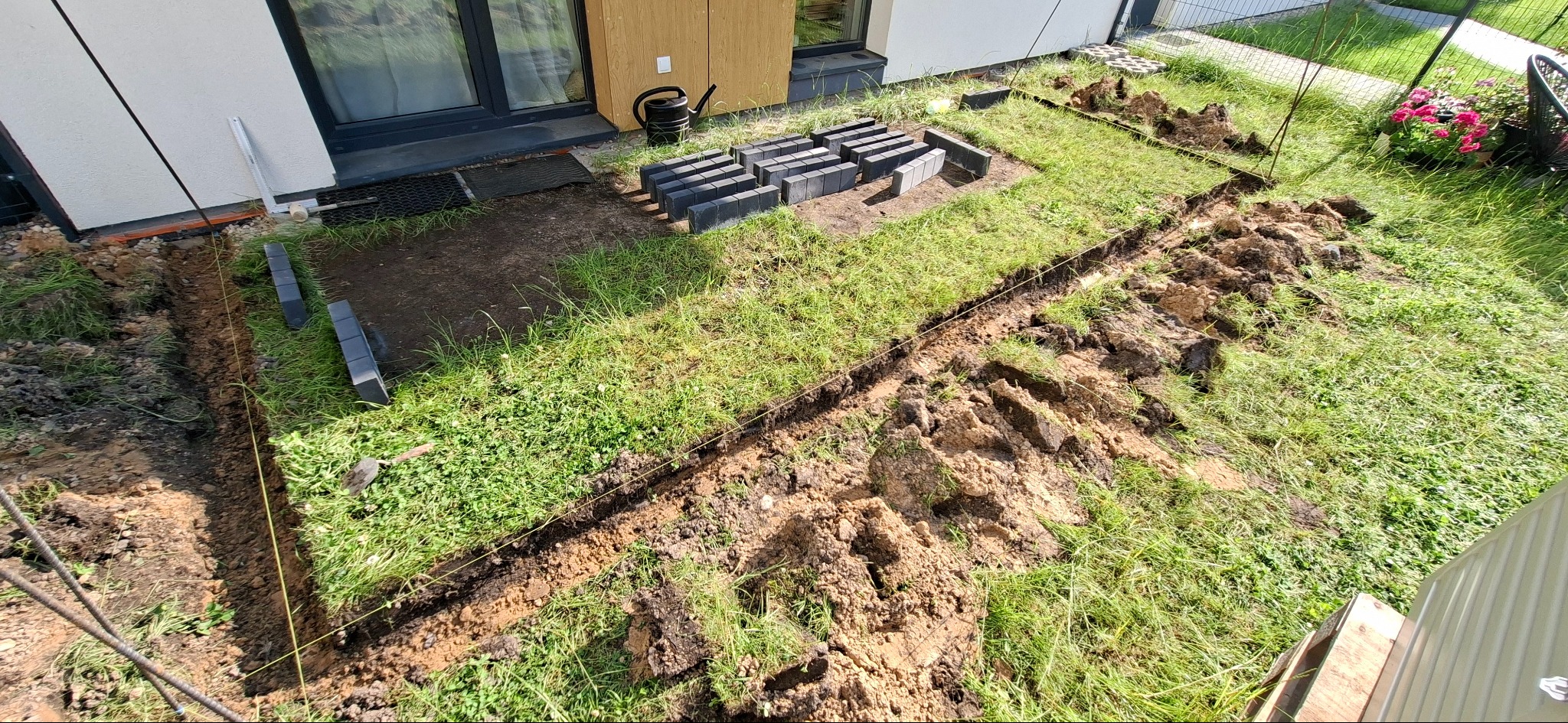 Przygotowanie terenu pod kostkę: wykopany rów, ułożone obrzeża z kostki, czarne kostki brukowe czekają na ułożenie obok domu z jasną elewacją.