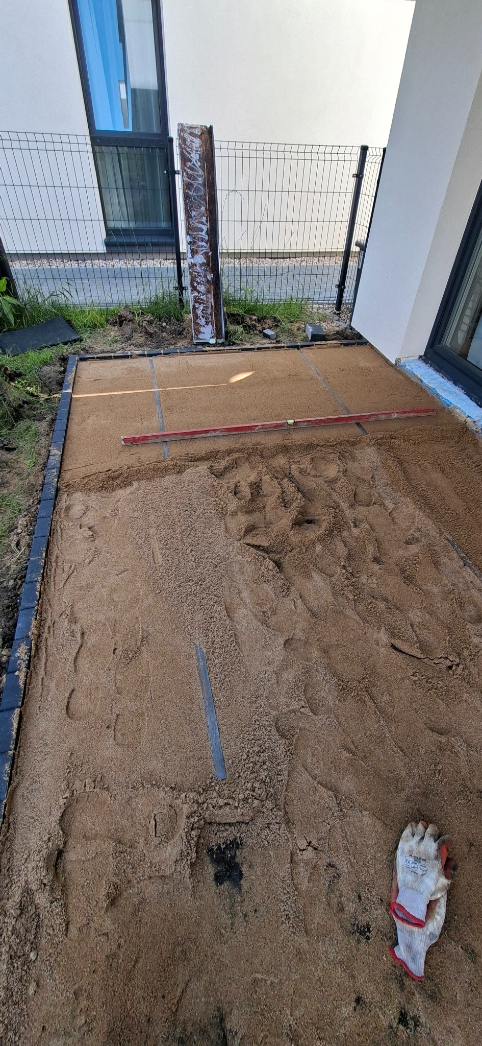 Przygotowanie podłoża pod kostkę brukową: wyrównany piasek, obrzeża, poziomica i rękawice robocze obok. Widoczne ślady stóp na piasku.