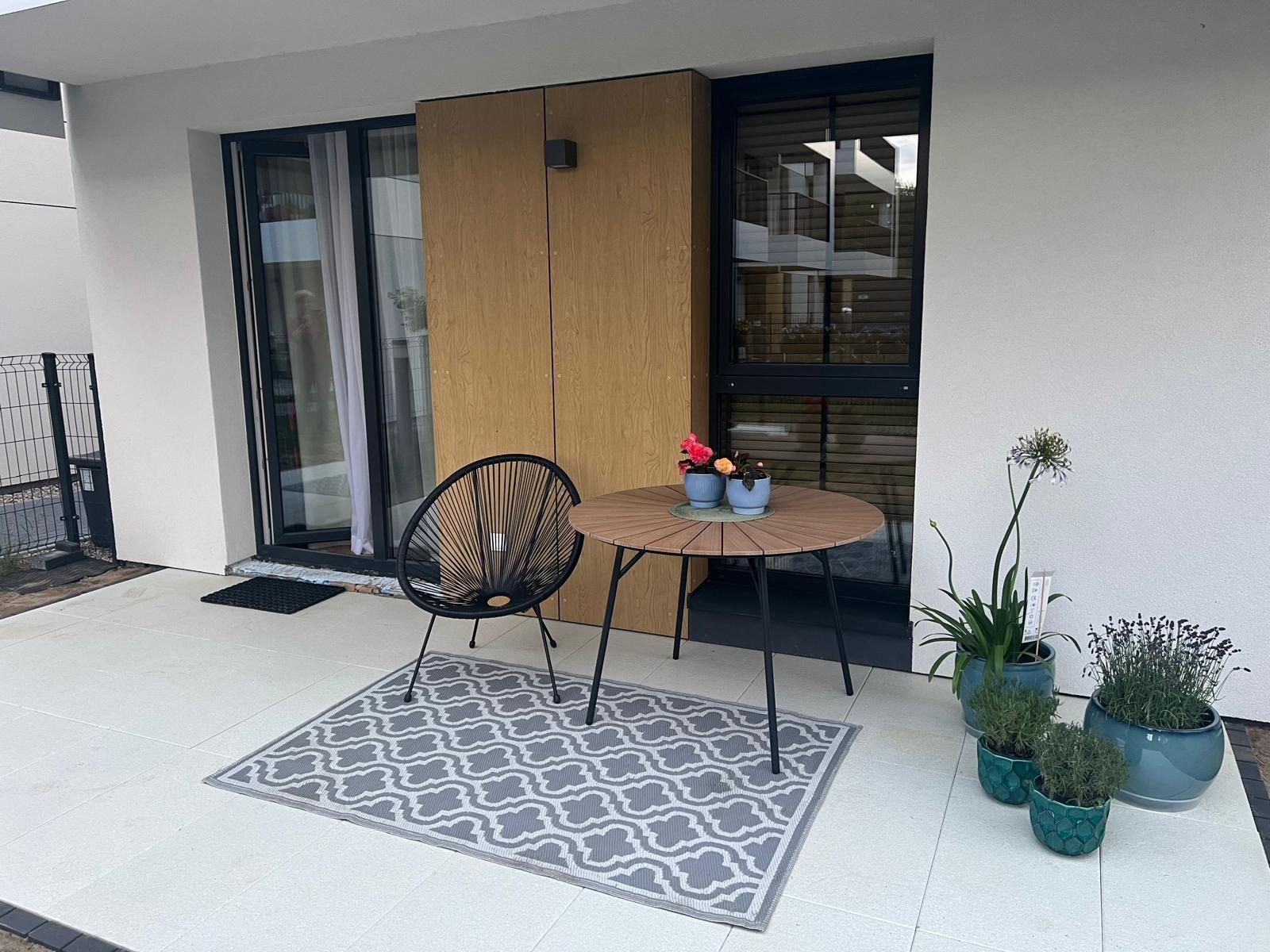 Biały taras z płyt, czarne krzesło, okrągły stolik z kwiatami, dywanik w szare wzory i rośliny w doniczkach tworzą przytulną strefę relaksu.