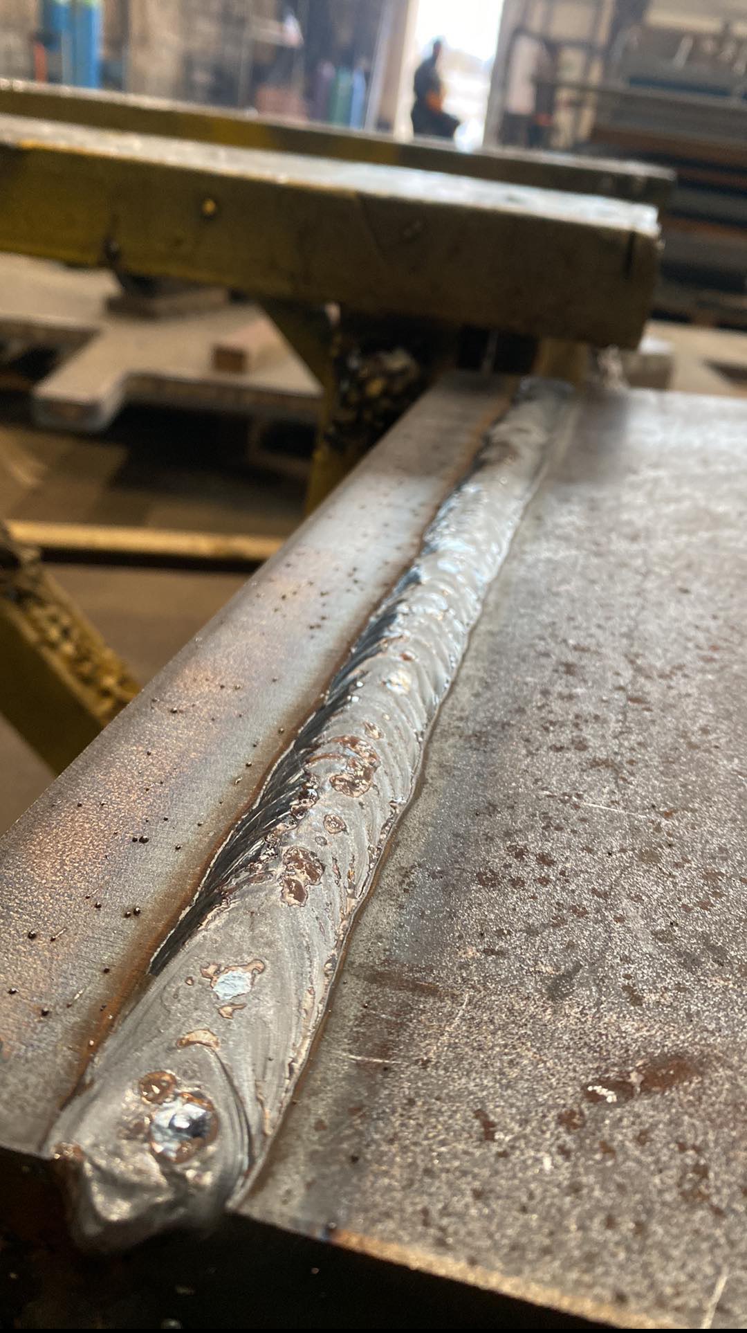 Świeżo wykonane złącze spawane na metalowej konstrukcji, widoczne ślady po spawaniu i drobne niedoskonałości. W tle hala produkcyjna i sylwetka pracownika.