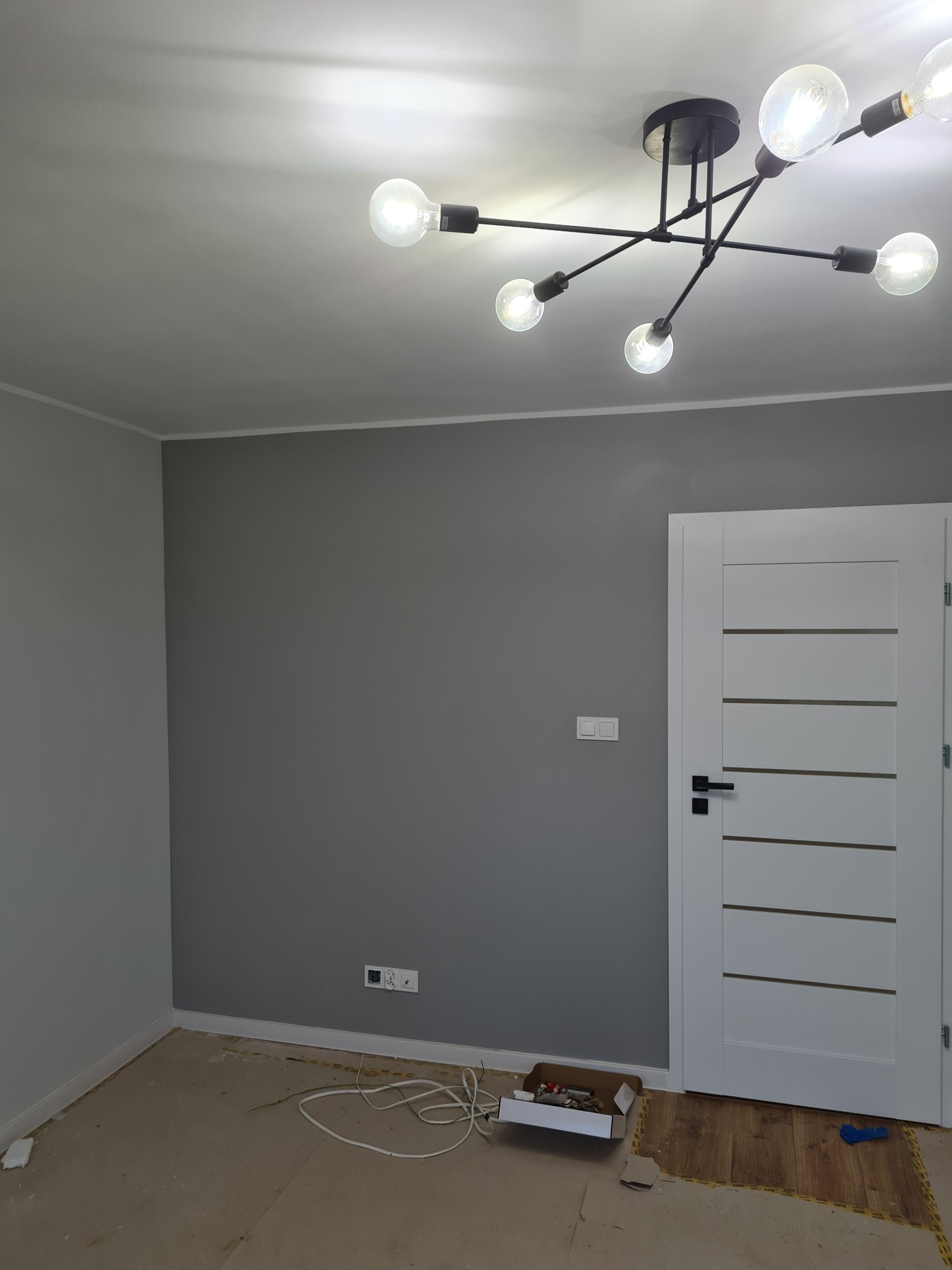 Szare ściany, białe drzwi z poziomymi frezami i nowoczesna lampa sufitowa w pokoju podczas prac wykończeniowych. Widoczne gniazdka i listwy przypodłogowe.