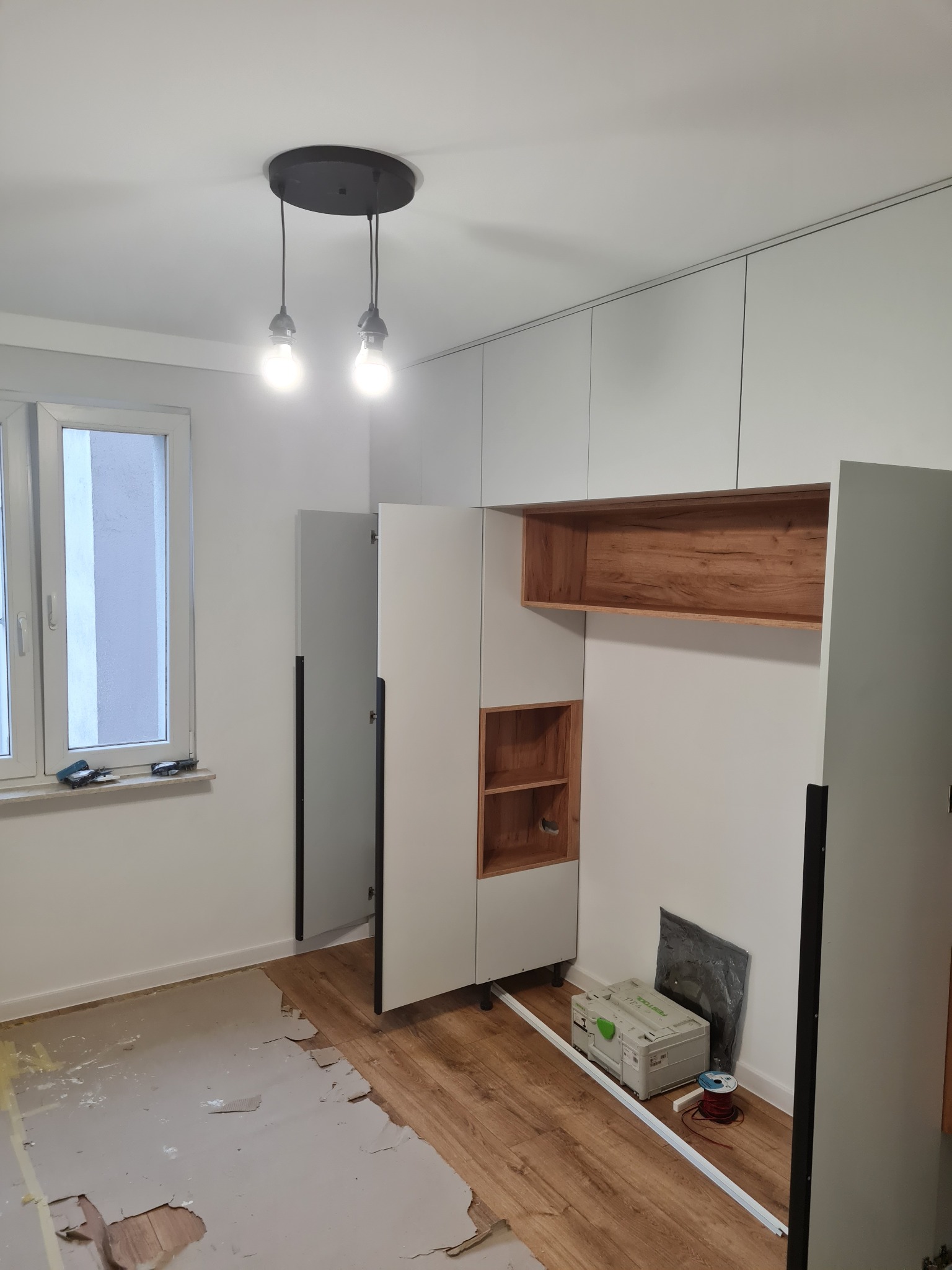Wnętrze w trakcie wykańczania: białe szafy w zabudowie, drewniane półki, podłoga częściowo wyłożona panelami, wisząca lampa z żarówkami. Widoczne narzędzia.