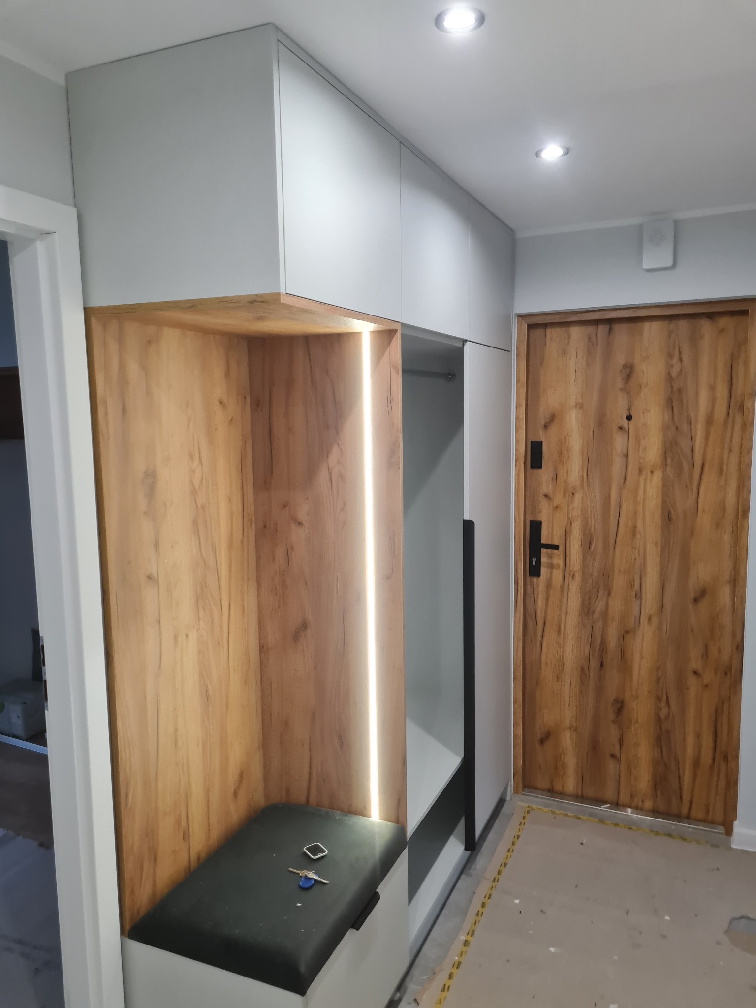 Nowoczesna zabudowa przedpokoju z drewnianą wnęką z oświetleniem LED i szarą szafką. Na siedzisku leżą klucze. Widoczna drewniana, brązowa drzwi.