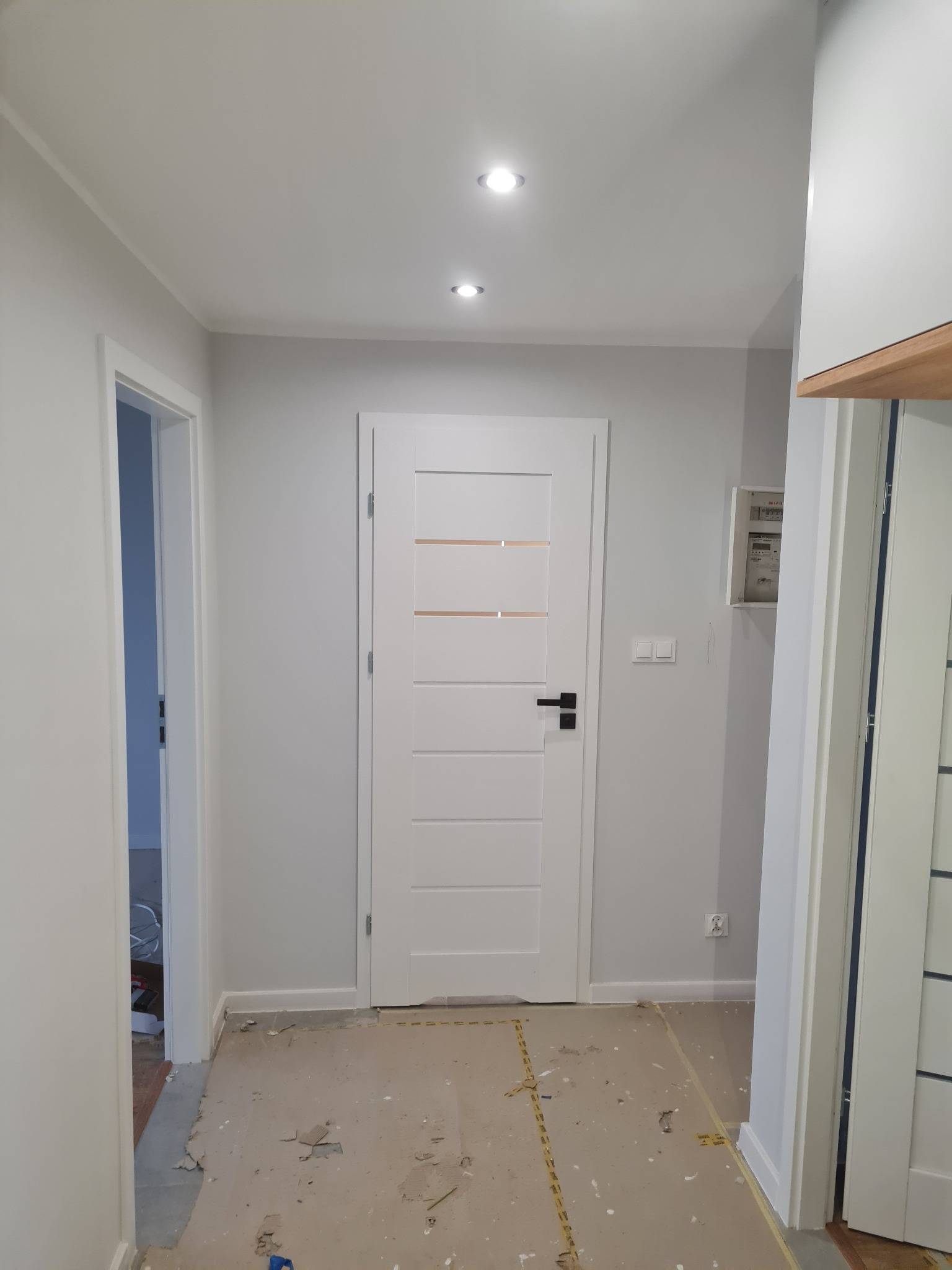 Jasne wnętrze w trakcie wykończenia: białe drzwi z przeszkleniami, szare ściany, zabezpieczona podłoga. Widoczne elementy instalacji elektrycznej.