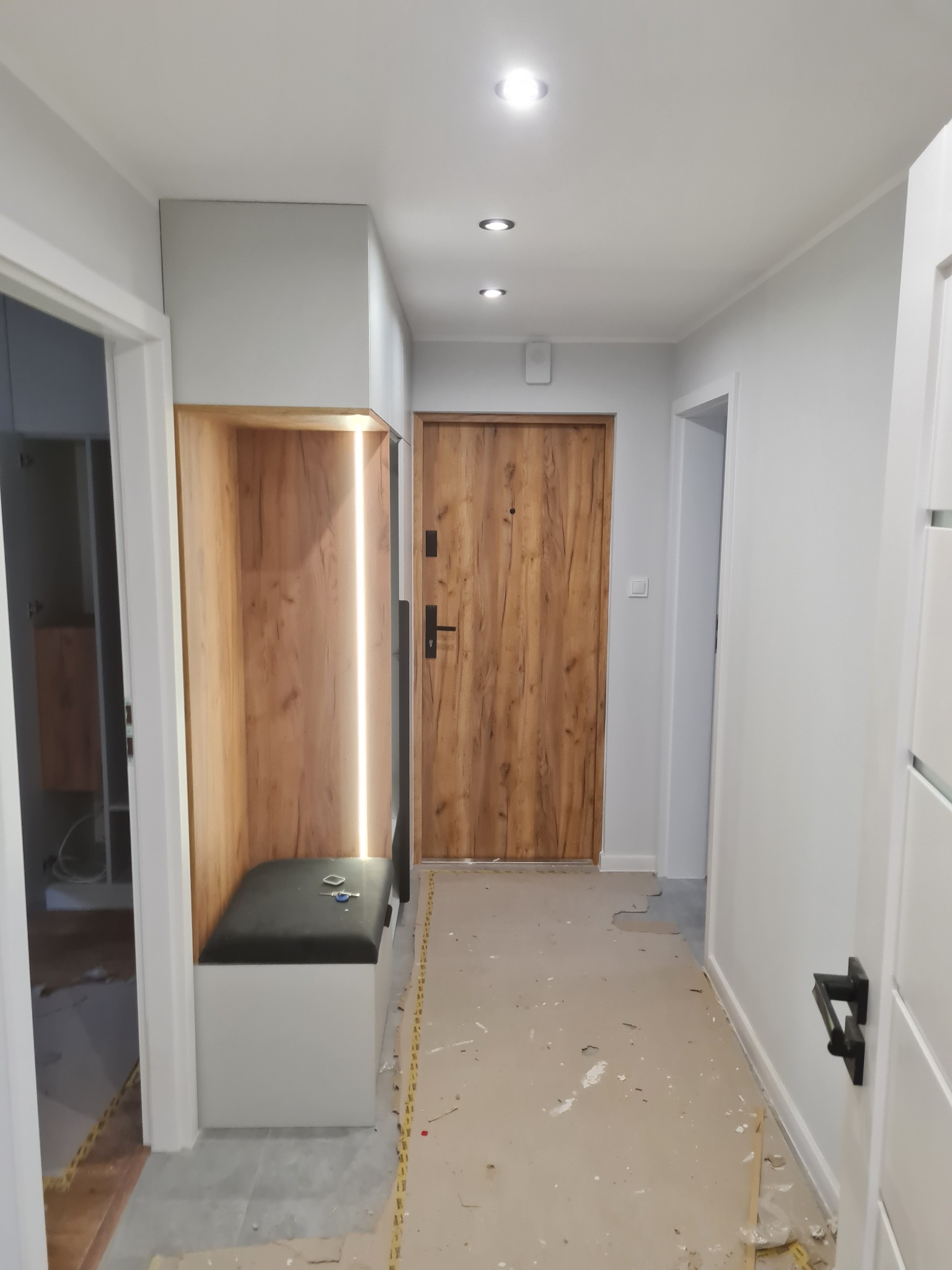 Nowoczesny przedpokój w trakcie wykończenia: drewniane drzwi wejściowe, zabudowa z siedziskiem i oświetleniem LED, białe ściany. Widoczne ślady prac remontowych na podłodze.