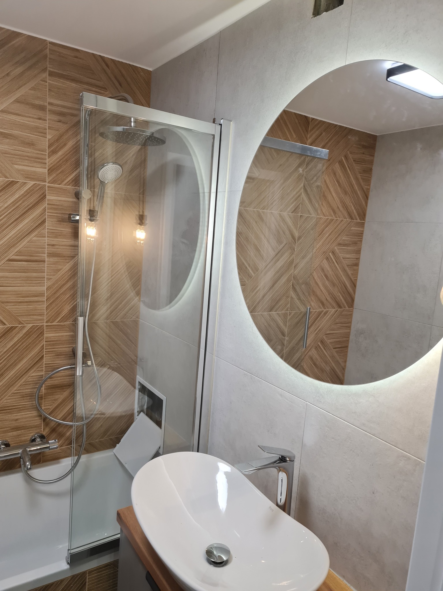 Nowoczesna łazienka z okrągłym lustrem LED, kabiną prysznicową ze szklanymi drzwiami i umywalką nablatową. Brązowe płytki z geometrycznym wzorem na ścianie.