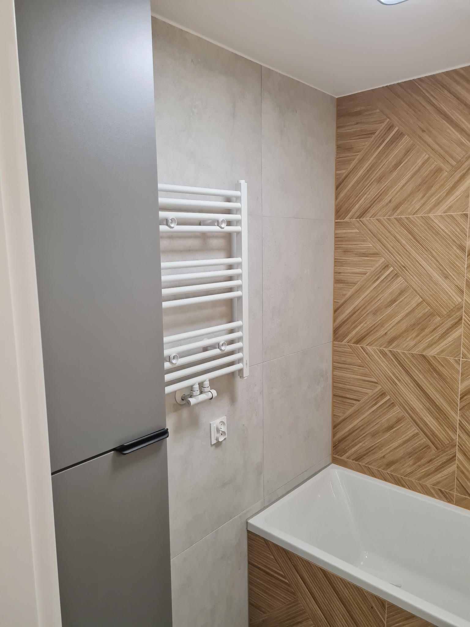 Nowoczesna łazienka z geometrycznym wzorem płytek na ścianie, grzejnikiem drabinkowym i szarą szafką. Widoczna wanna i gniazdko elektryczne.