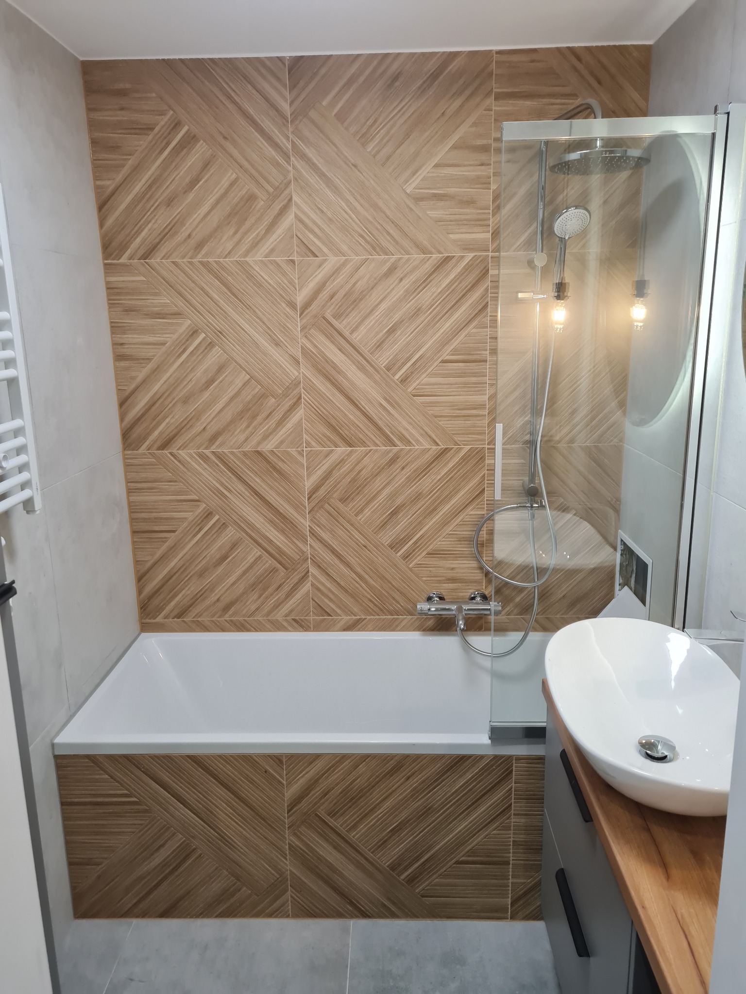 Nowoczesna łazienka z wanną, szklanym parawanem i umywalką nablatową. Płytki w geometryczny wzór imitują drewno, tworząc ciepły kontrast z szarością i bielą.