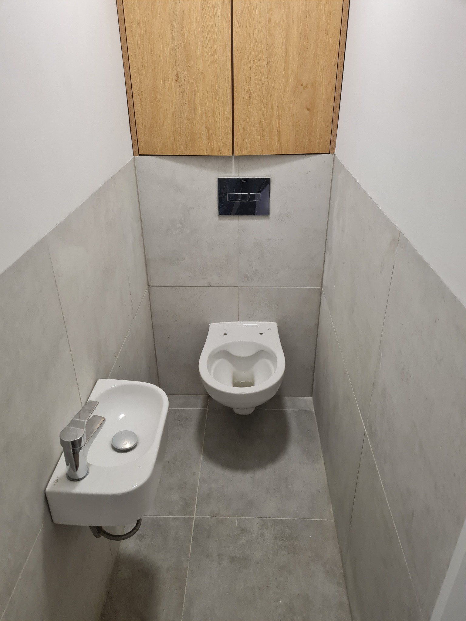 Nowoczesna, minimalistyczna toaleta z wiszącą muszlą, umywalką i szafką. Szare płytki na ścianach i podłodze. Czarny panel sterujący spłuczką.