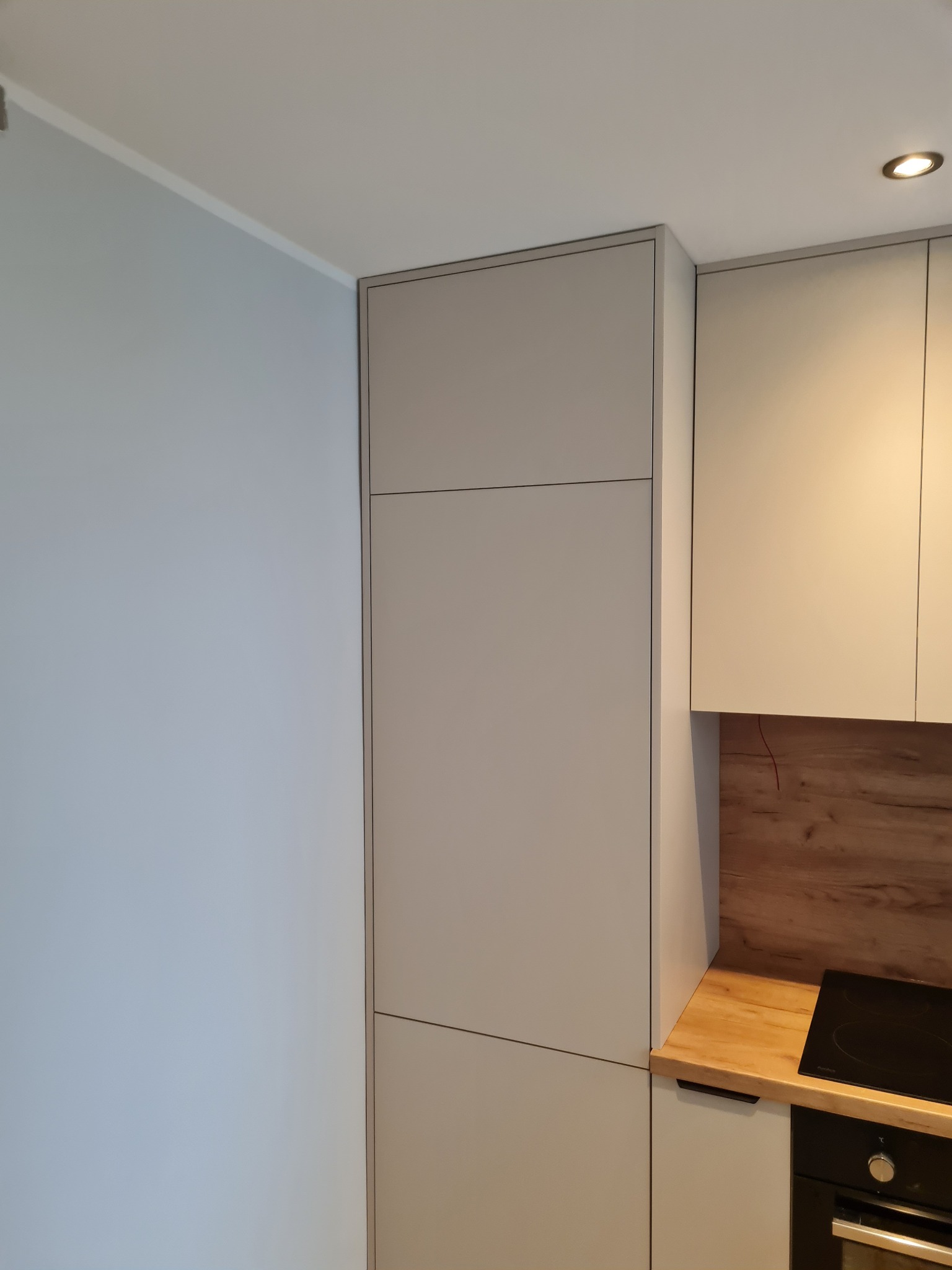 Minimalistyczna zabudowa kuchenna w odcieniach szarości i drewna. Widoczne szafki, blat i płyta indukcyjna. Oświetlenie punktowe w suficie.