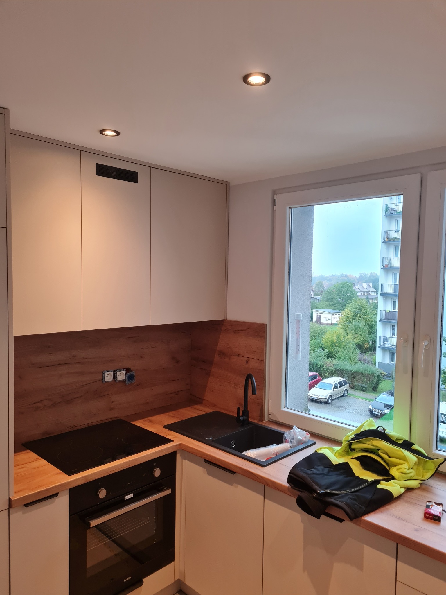 Nowoczesna kuchnia z drewnianym blatem i białymi szafkami. Widoczne okno z widokiem na zewnątrz i czarny zlew. Na blacie leży kurtka robocza i paczka papierosów.