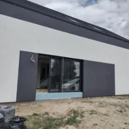 Nowoczesny dom z elewacją w odcieniach szarości i bieli, widoczne okno przesuwne i materiały budowlane na ziemi. Minimalistyczny design budynku.