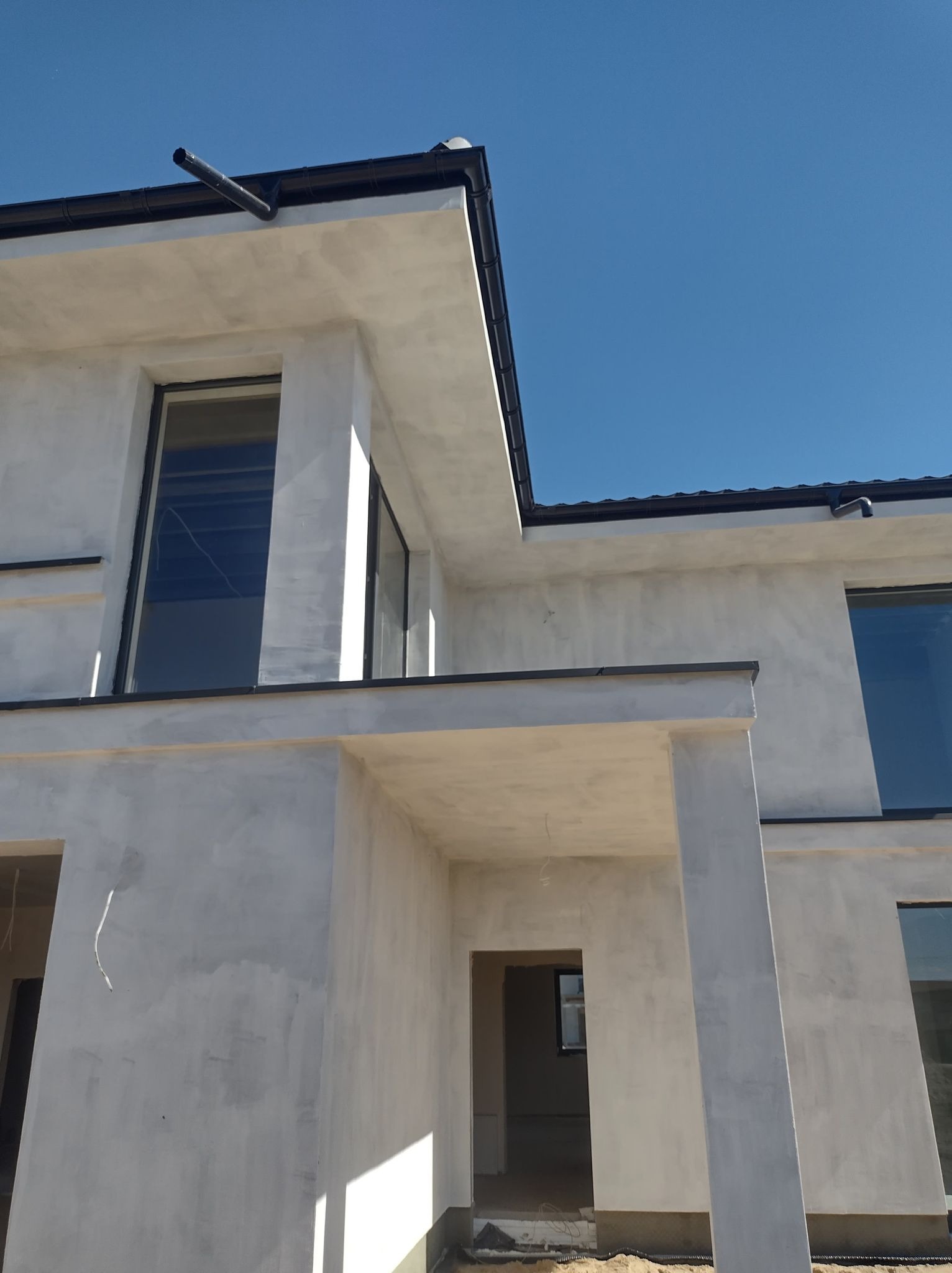 Surowy, dwukondygnacyjny dom z szarą elewacją w trakcie wykańczania, z widocznymi oknami i elementami dachu na tle błękitnego nieba. Ujęcie z dołu.