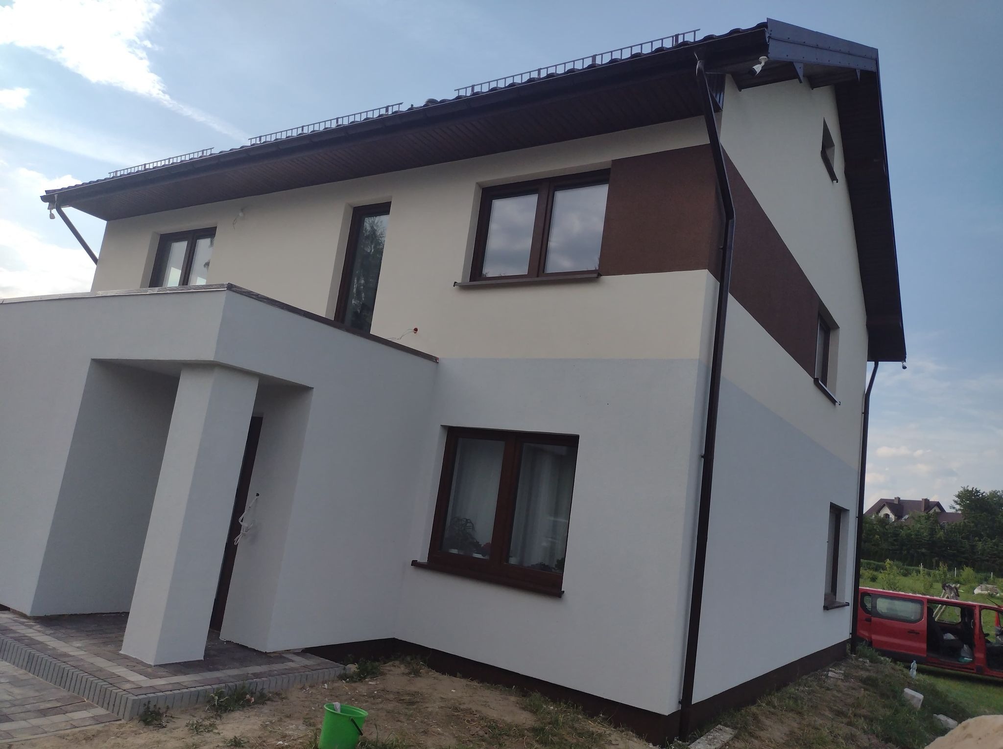 Dwukondygnacyjny dom po renowacji elewacji, z kremową górną częścią i szarą dolną, brązowe akcenty, widoczne okna i fragment czerwonego samochodu.