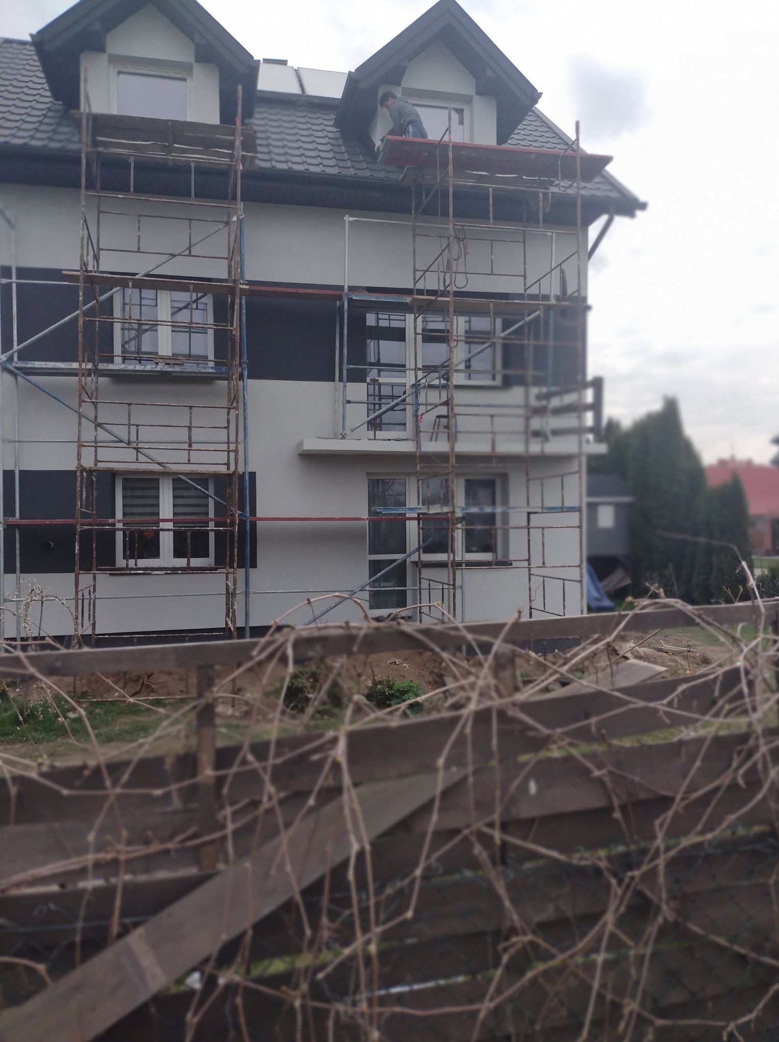 Dom w rusztowaniach, świeżo otynkowany, z elementami w kolorze grafitowym. Pracownik na górze. Widok zza drewnianego płotu.