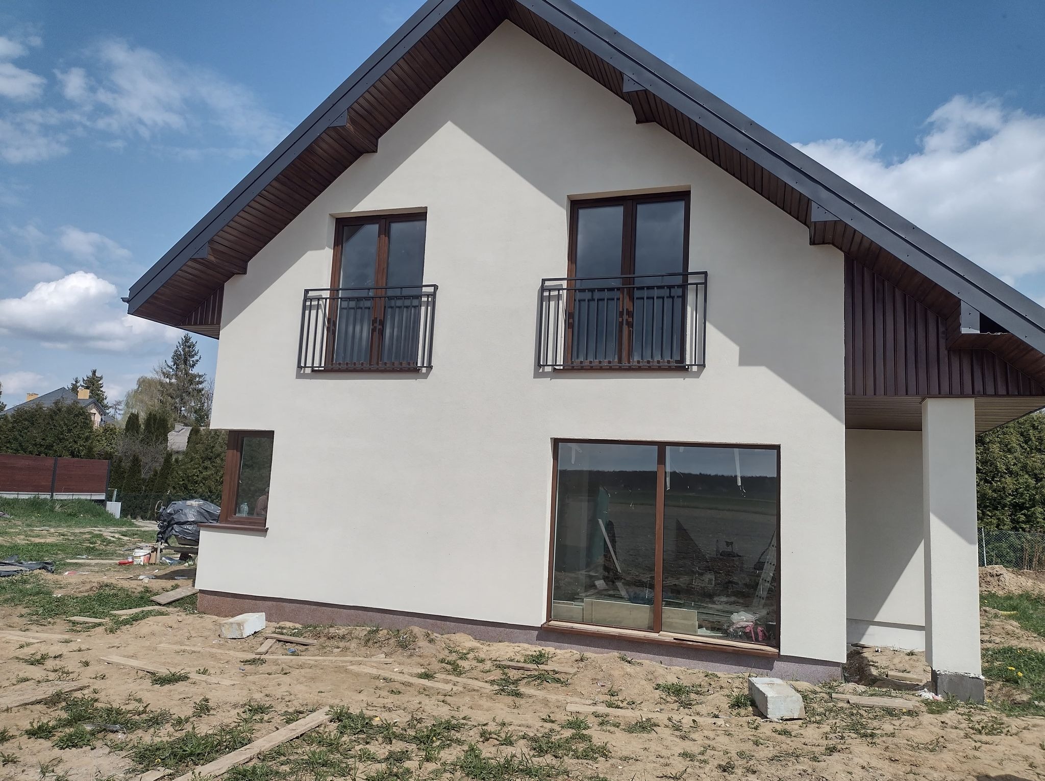 Dwukondygnacyjny dom z jasną elewacją, ciemną stolarką okienną i drewnianą podbitką dachową. Widoczne balkony z czarną, metalową balustradą oraz fragment niezagospodarowanego terenu.