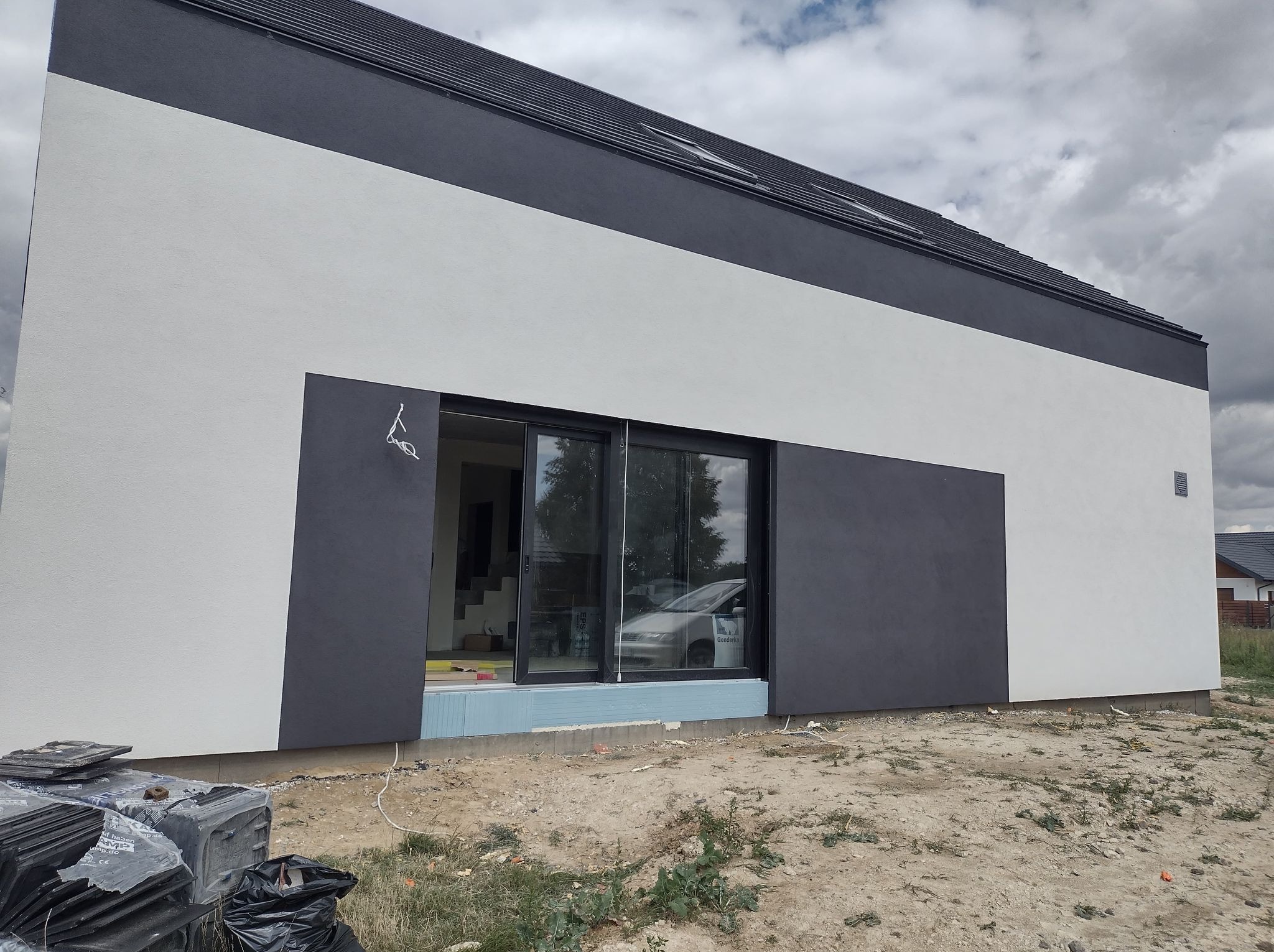 Nowoczesny dom z elewacją w odcieniach szarości i bieli, widoczne okno przesuwne i materiały budowlane na ziemi. Minimalistyczny design budynku.