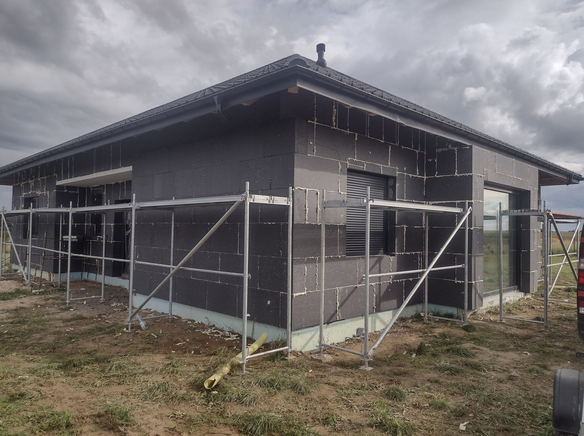 Dom w Kaznów-Kolonia w trakcie ocieplania. Widoczne rusztowania i czarne płyty izolacyjne na ścianach. Etap prac elewacyjnych, szare niebo w tle.