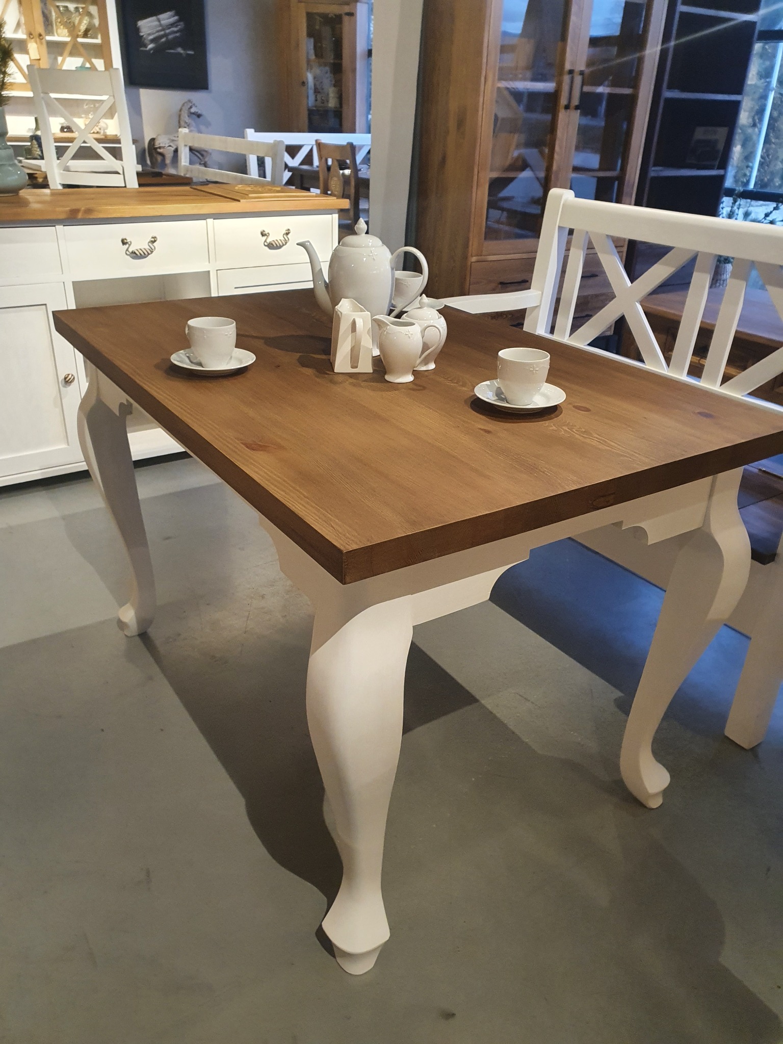 Drewniany stół z białymi, rzeźbionymi nogami, nakryty do herbaty. W tle kredens i krzesła w stylu prowansalskim, tworzące spójną aranżację wnętrza.