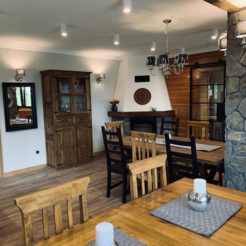 Rustykalna jadalnia z drewnianym stołem, krzesłami i kredensem. Kominek z kamienia i drewna. Elementy dekoracyjne w stylu wiejskim. Oświetlenie punktowe.