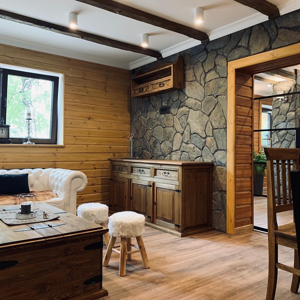 Rustykalne wnętrze: drewniana komoda z szufladami i szafkami, kamienna ściana, drewniane belki stropowe, biała sofa i pufy. Widok na sąsiednie pomieszczenie.