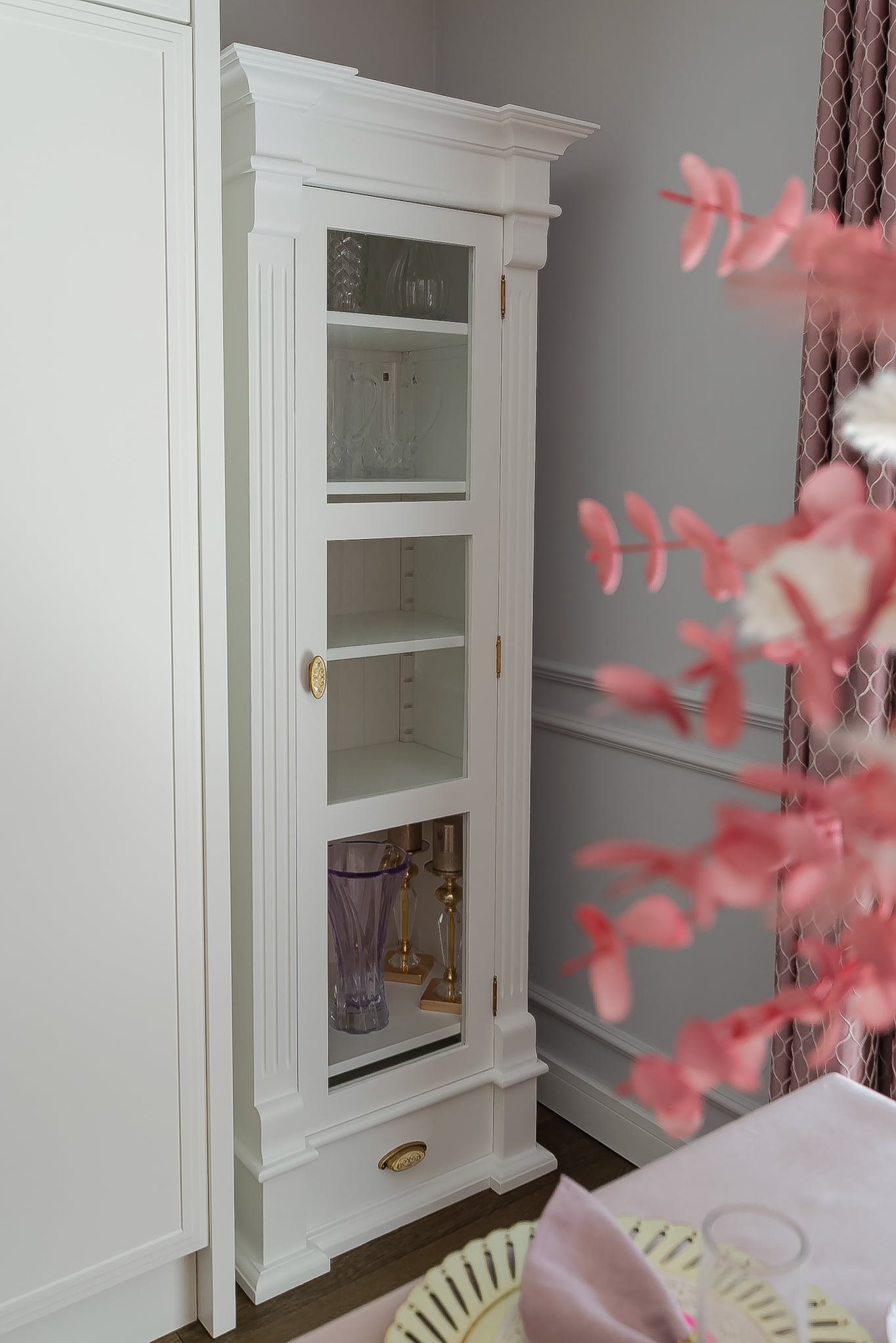 Biała, przeszklona witryna w stylu klasycznym, z dekoracyjnymi listwami i złotymi uchwytami, stojąca obok białej szafy, na tle szarej ściany.