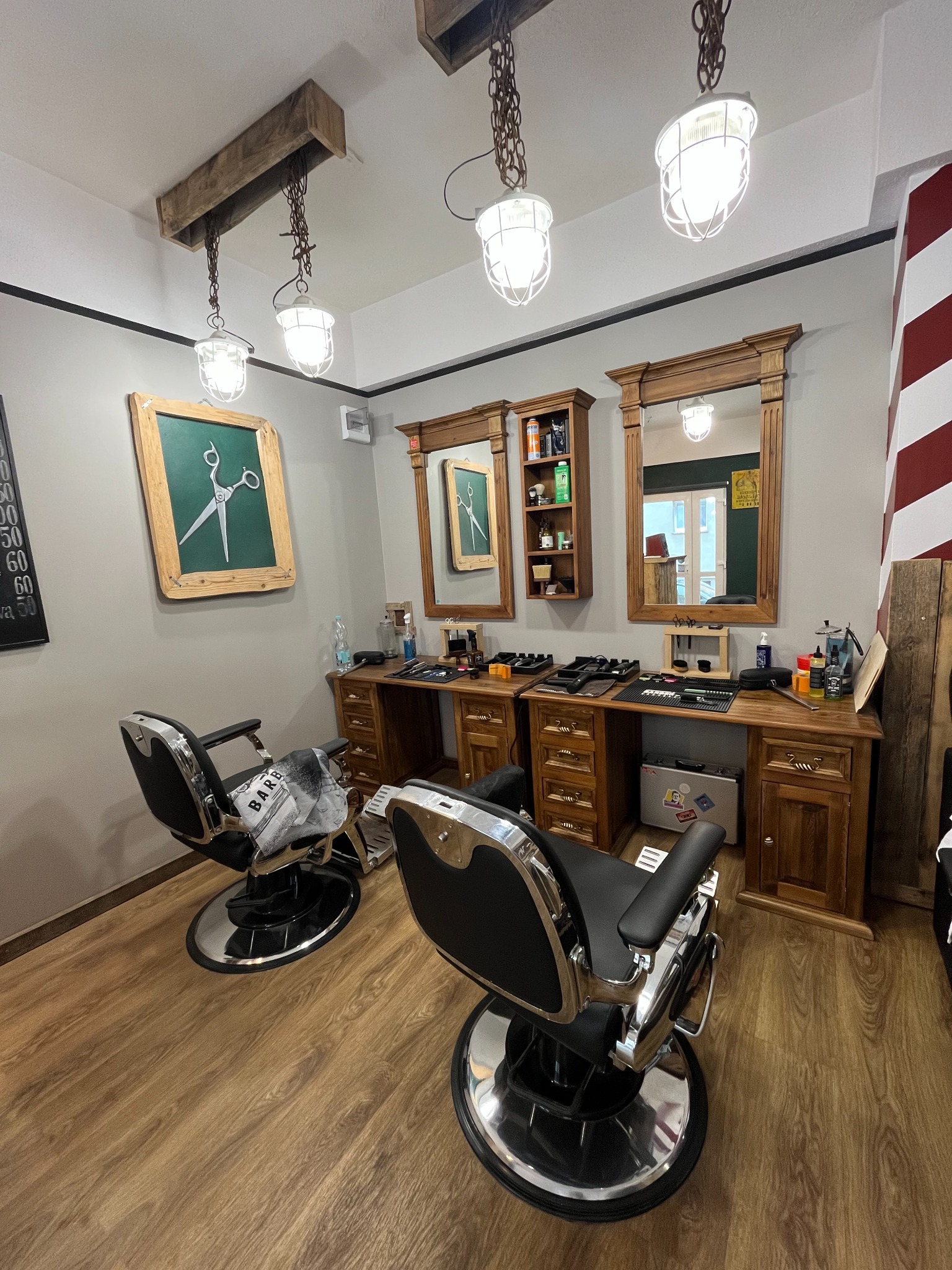 Wnętrze salonu barberskiego z dwoma stanowiskami: drewniane konsole z lustrami, chromowane fotele, dekoracje w stylu retro i oświetlenie industrialne.
