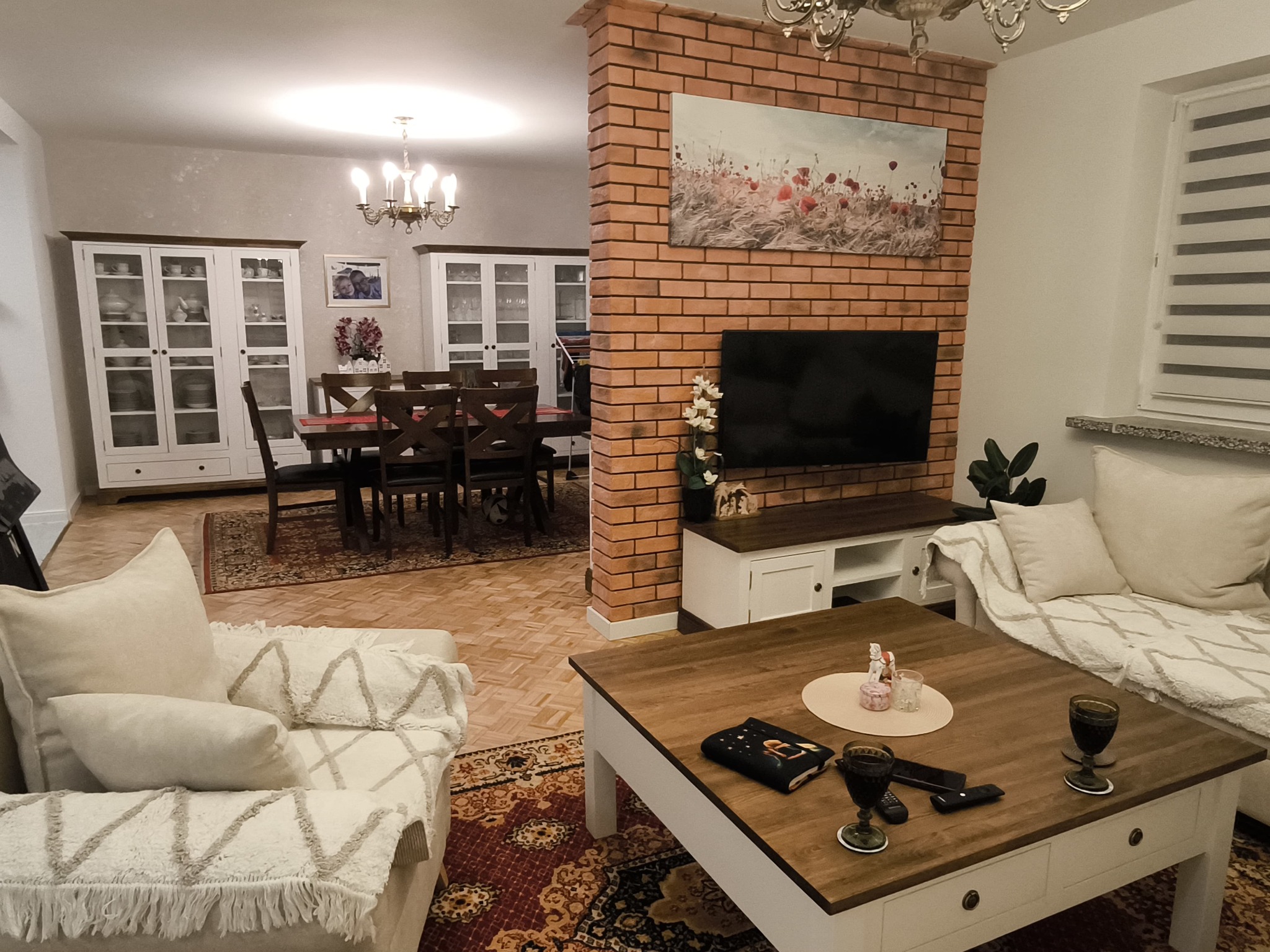 Salon z ceglaną ścianą, drewnianym stołem i białymi szafkami. Widoczne elementy dekoracyjne i oświetlenie, tworzące przytulną atmosferę wnętrza.