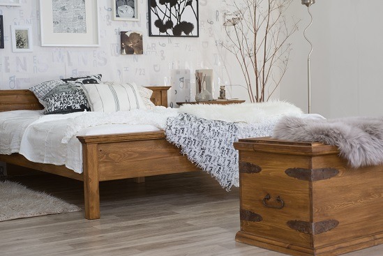 Drewniane łóżko z skrzynią w rustykalnym stylu, dekorowane poduszkami i pledem, na tle ściany z wzorem liter i galerii obrazów. Oświetlenie dzienne.