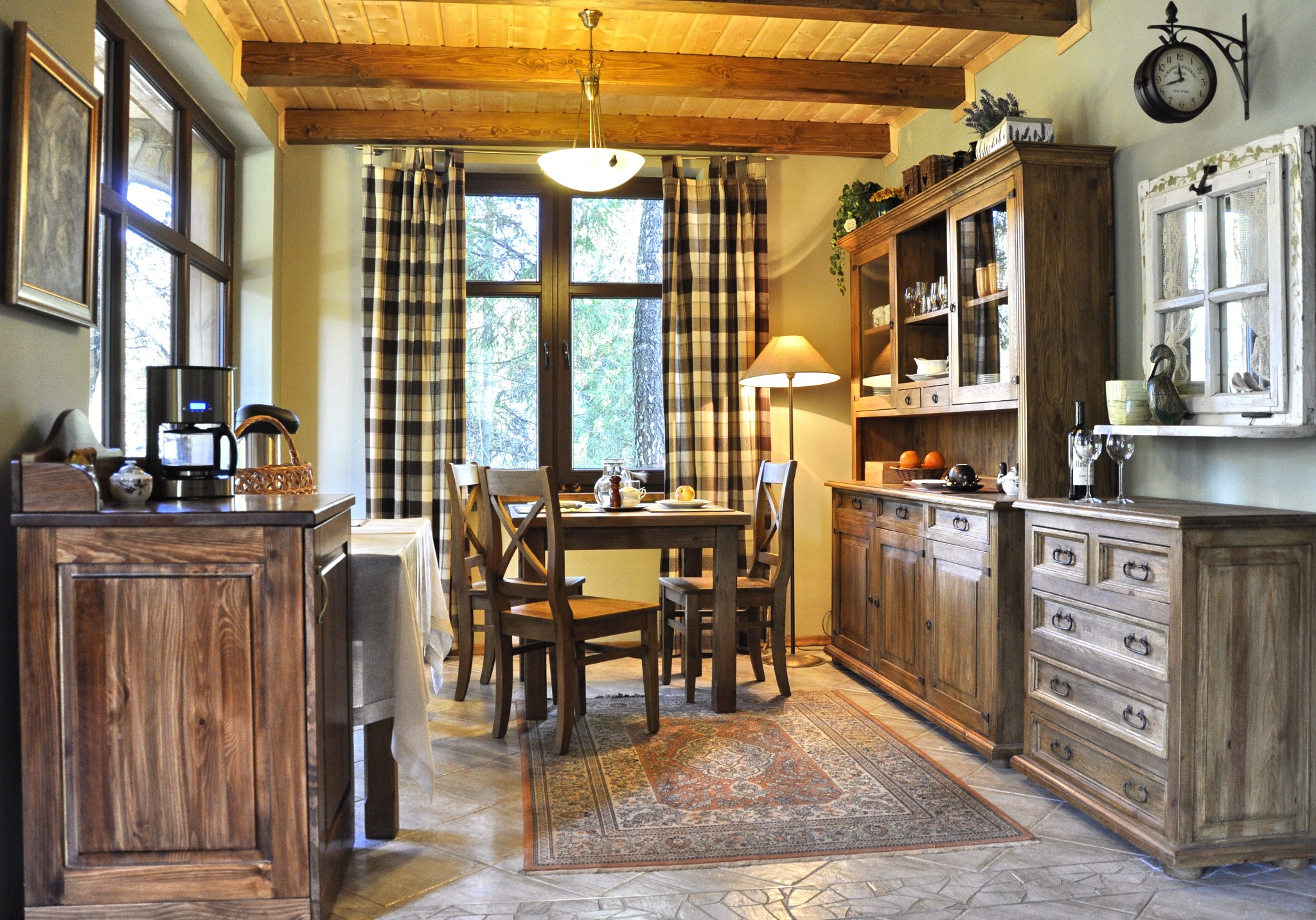 Rustykalna jadalnia z drewnianymi meblami: kredens, stół z krzesłami, komoda. Okno z kraciastymi zasłonami, lampa i dywan dopełniają wystrój.