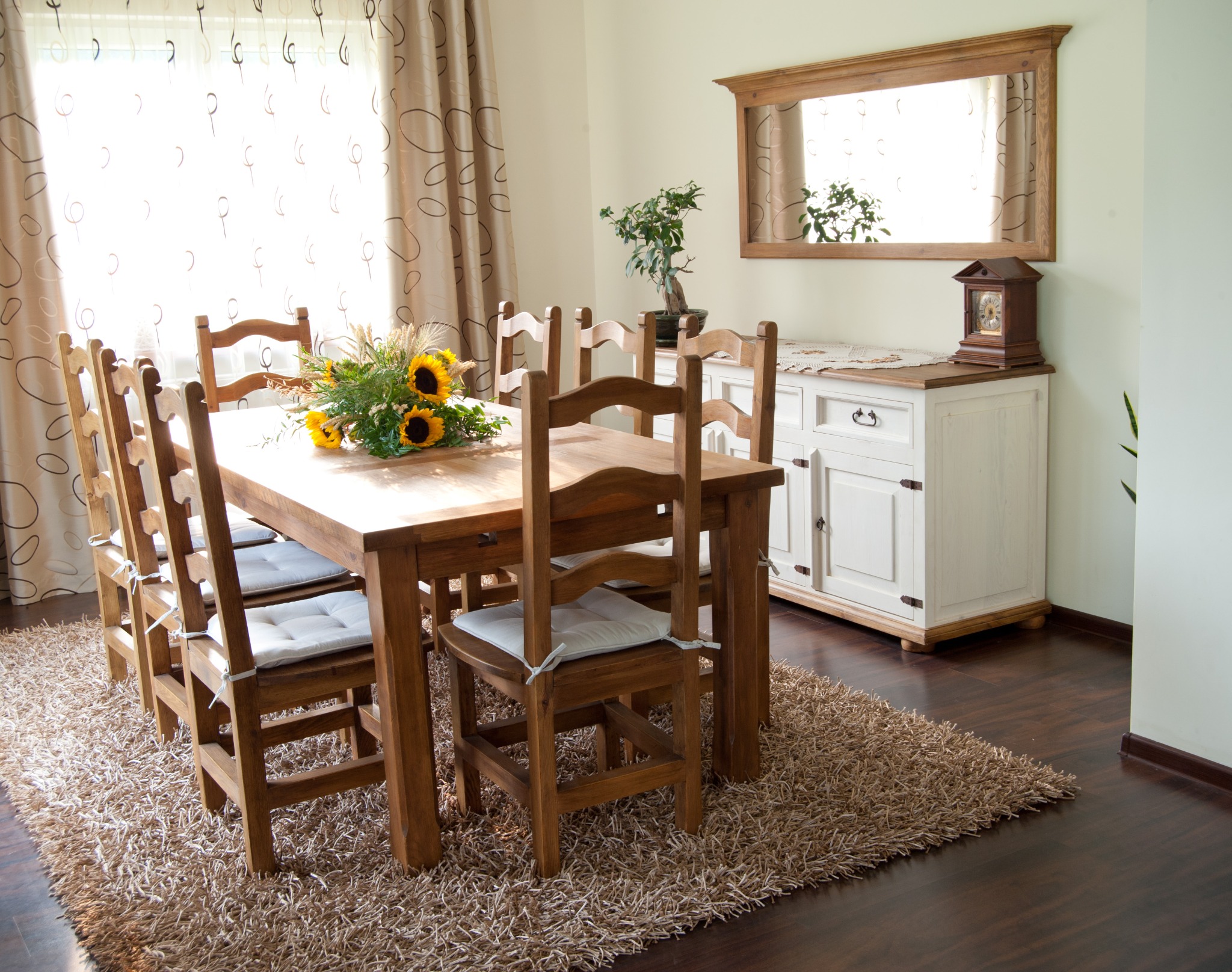 Drewniany stół z krzesłami, bukiet słoneczników, komoda z lustrem w ramie, parkiet i dywan tworzą przytulną jadalnię w jasnym wnętrzu domu.