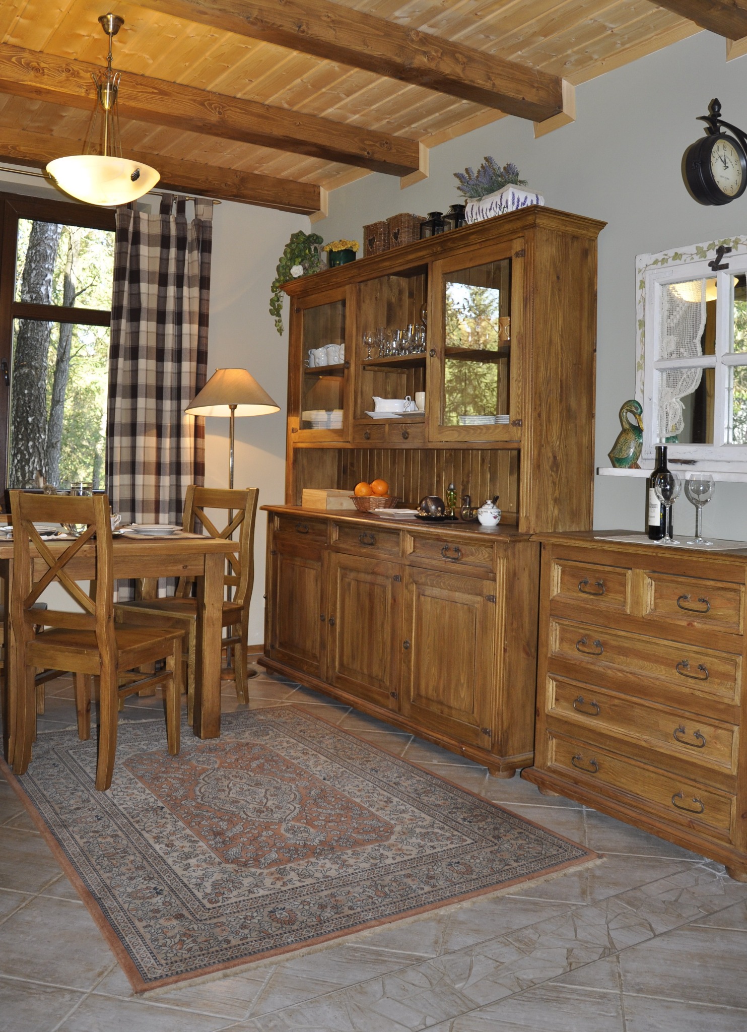 Rustykalna jadalnia z drewnianym stołem, krzesłami i kredensem. Na kredensie dekoracje, a na stole nakrycia. Okno z widokiem na zieleń. Drewniany sufit z belkami.