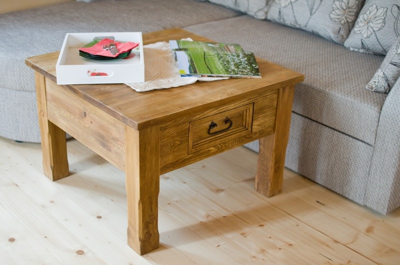 Drewniany stolik kawowy z szufladą i metalowym uchwytem, na blacie taca z naczyniami i czasopisma, obok sofa z poduszkami w kwiaty, parkiet z jasnego drewna.