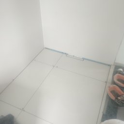 Ogólnobudowlane - Układanie jasnych płytek podłogowych w łazience, widoczna listwa przypodłogowa i narzędzia glazurnicze. Etap prac z zachowaniem dystansu między płytkami.