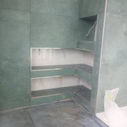 Ogólnobudowlane - Narożna zabudowa z płyt GK w surowym stanie, z widocznymi przewodami elektrycznymi. Szare płyty na ścianach i podłodze, białe wnętrze wnęk.