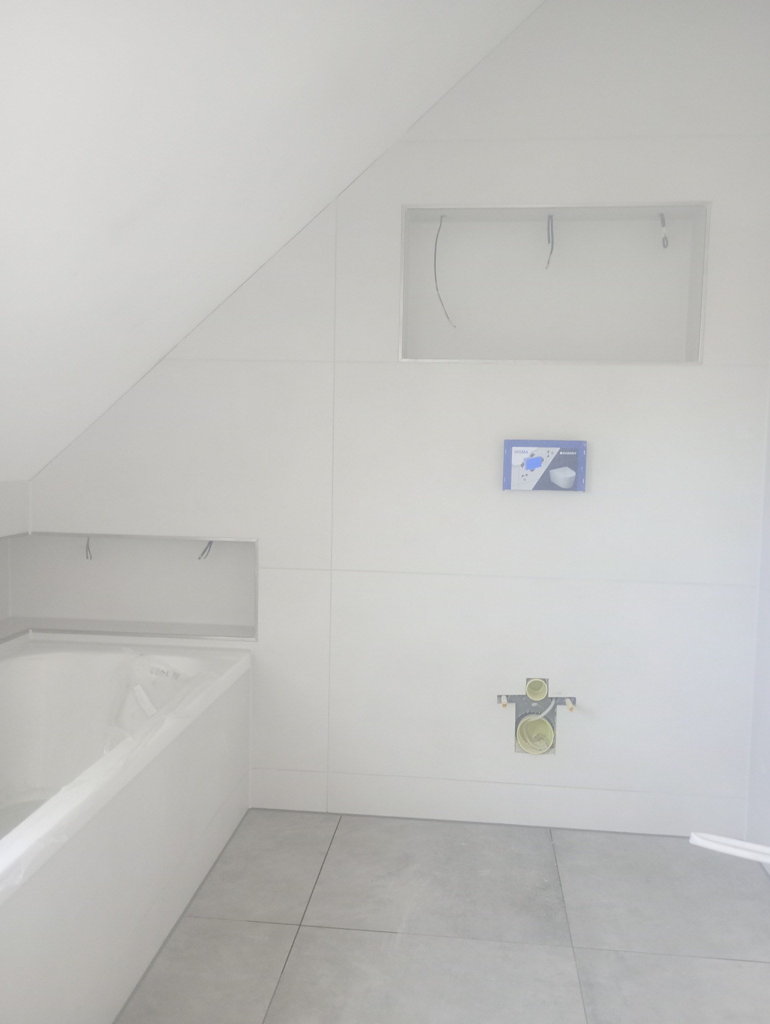 Biała łazienka w trakcie wykańczania: obudowana wanna, ułożone szare płytki podłogowe, przygotowane instalacje, wnęki w ścianach, zabudowa na poddaszu.