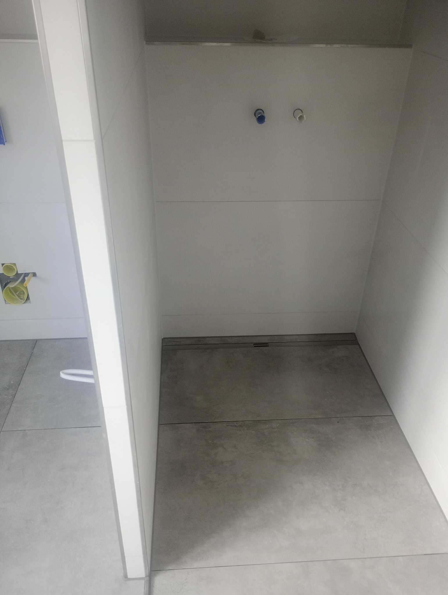 Minimalistyczna kabina prysznicowa w trakcie wykańczania, widoczne szare płytki podłogowe, białe ściany i przygotowane przyłącza wodne.