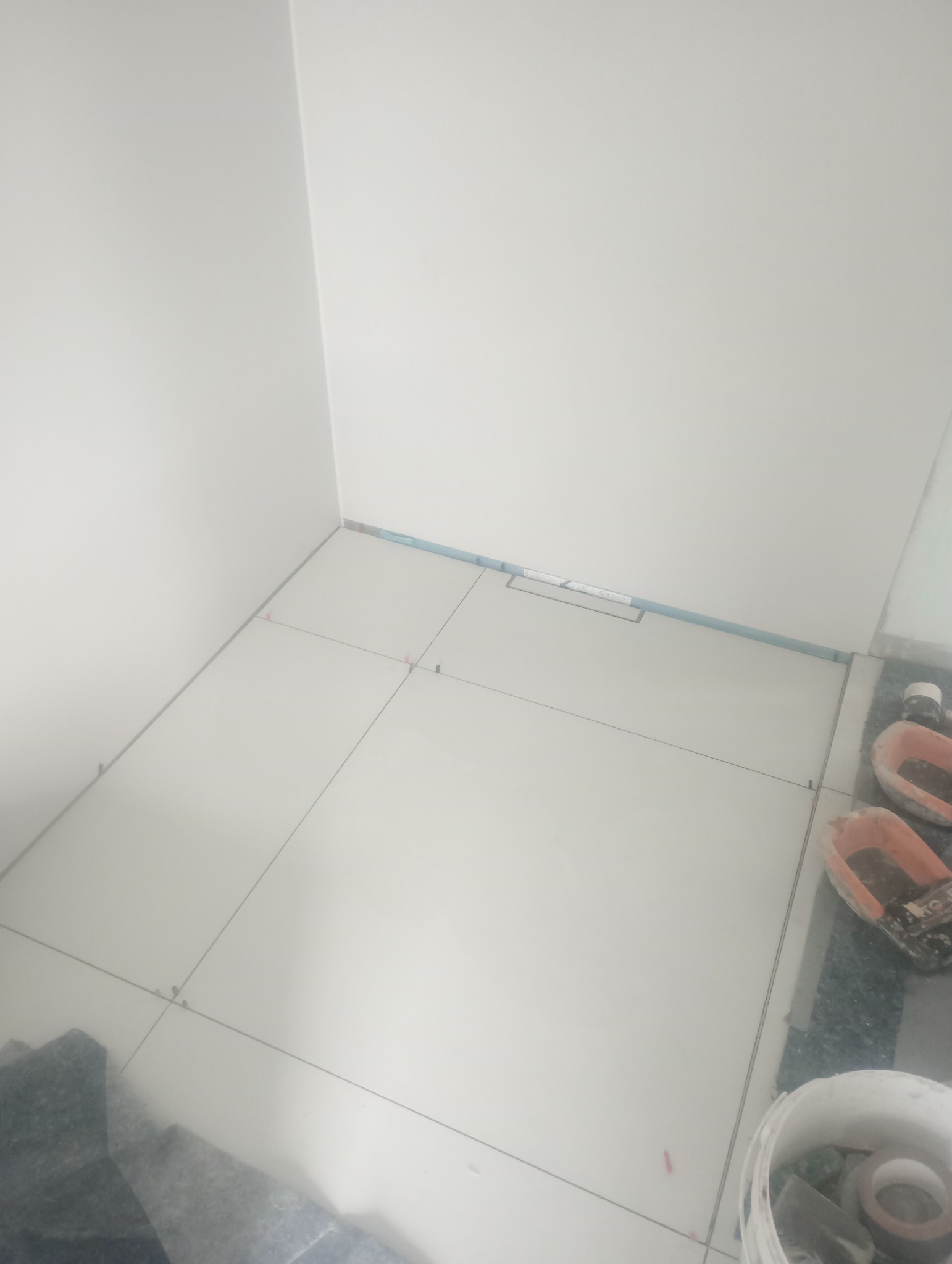 Układanie jasnych płytek podłogowych w łazience, widoczna listwa przypodłogowa i narzędzia glazurnicze. Etap prac z zachowaniem dystansu między płytkami.