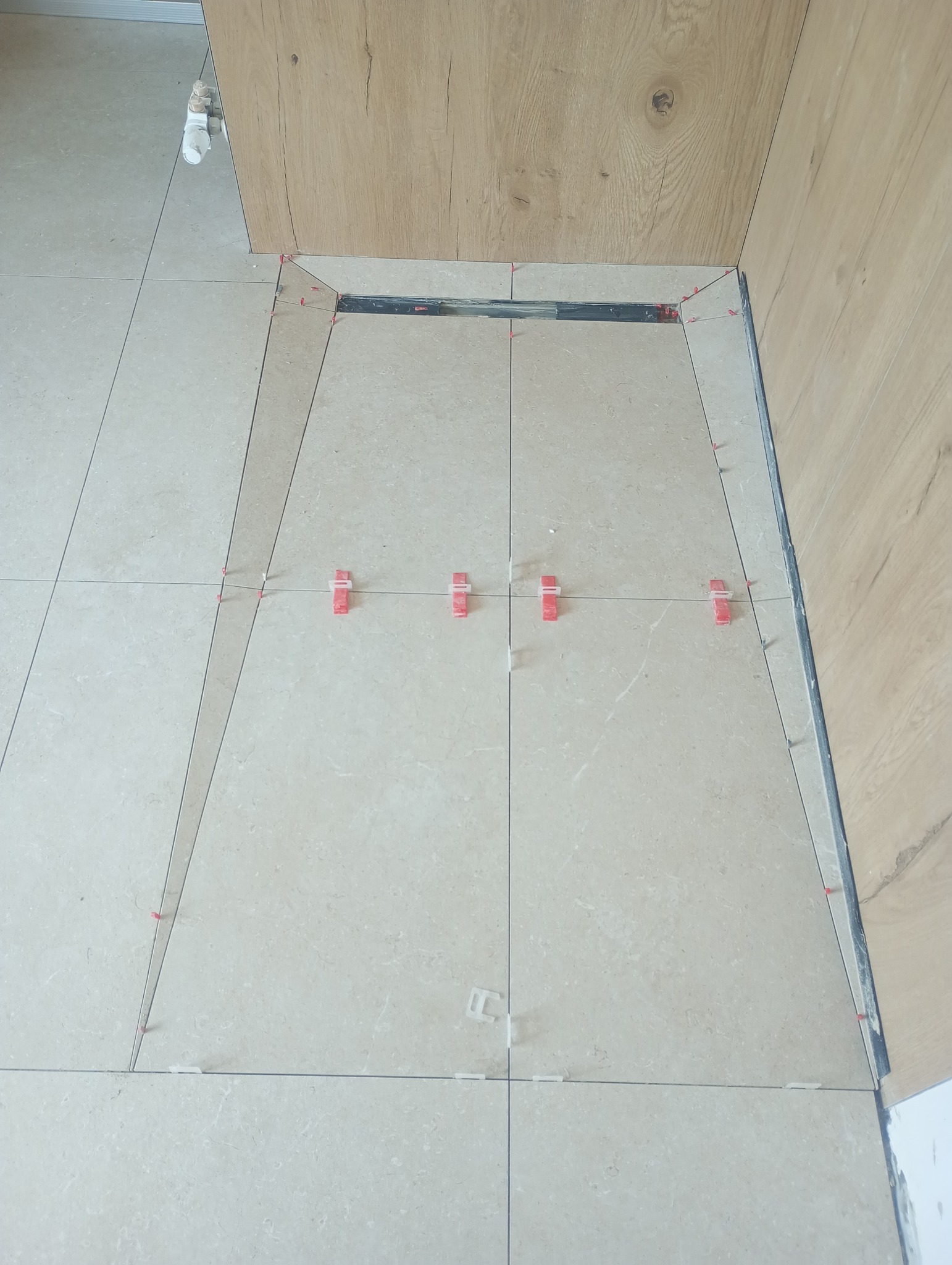 Beżowe płytki podłogowe, ułożone wokół odpływu liniowego, z użyciem czerwonych i białych klinów poziomujących. Widoczne fragmenty drewnopodobnej zabudowy.