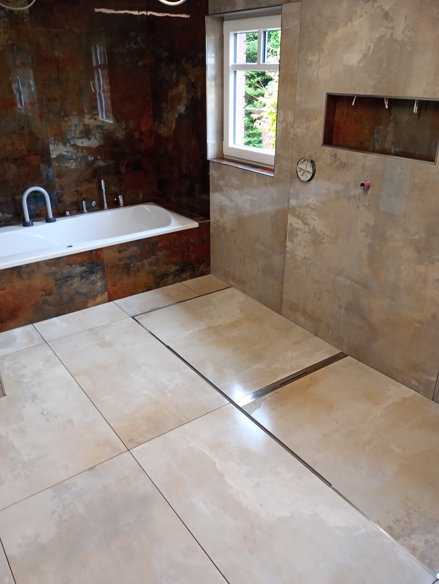 Nowoczesna łazienka z wanną, beżowymi płytkami podłogowymi i ściennymi imitującymi kamień. Widoczne odpływy liniowe i okno z widokiem na zieleń.