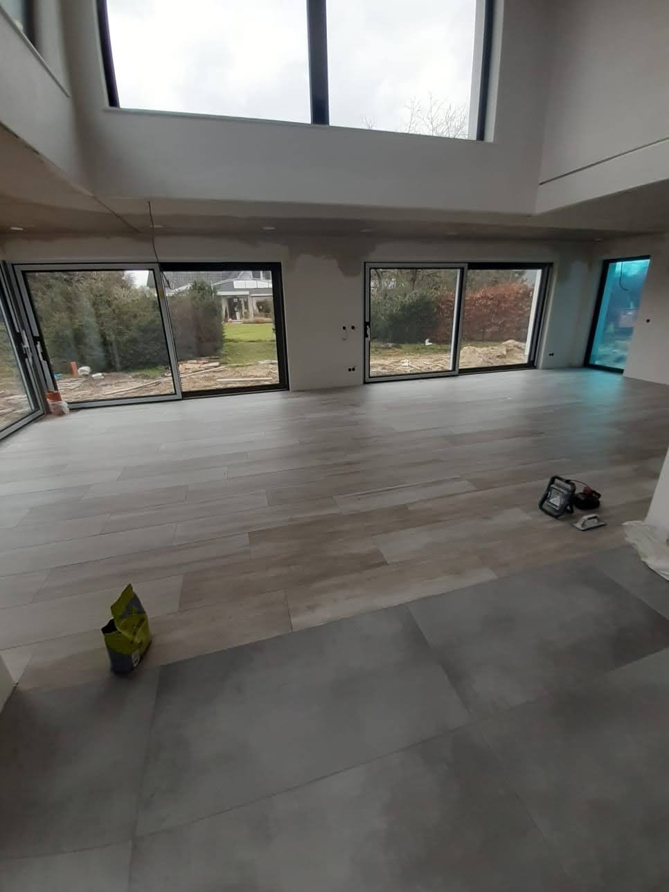 Jasne płytki podłogowe w przestronnym wnętrzu z dużymi oknami i widokiem na ogród. Częściowo wykończone ściany, narzędzia glazurnicze w tle.