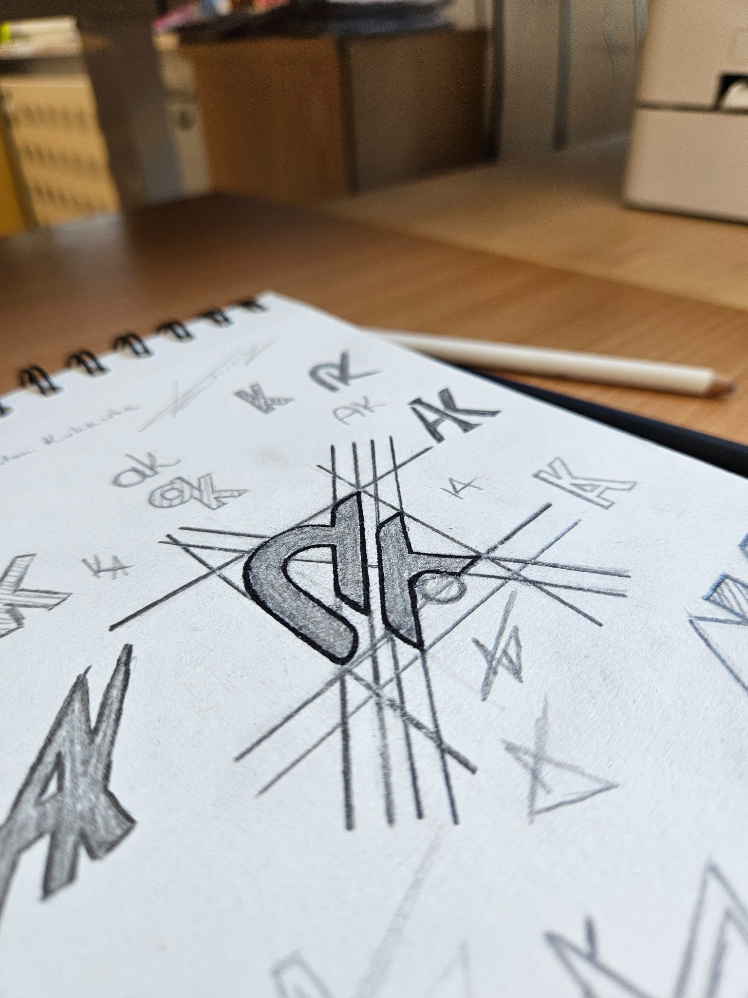 Szkicownik z koncepcjami logo 'AK' w różnych wariantach graficznych, wykonany ołówkiem. Widoczne linie pomocnicze i notatki. Kadr z bliska, perspektywa z góry.