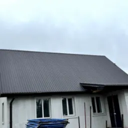 Szary dach z blachy trapezowej na nowym domu z białymi ścianami. Widoczne rusztowania i okna. Pochmurne niebo w tle.