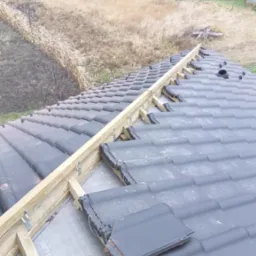 Montaż dachówki ceramicznej na drewnianej konstrukcji z folią w tle wiejskiego krajobrazu. Widok z góry na fragment dachu w trakcie prac dekarskich.