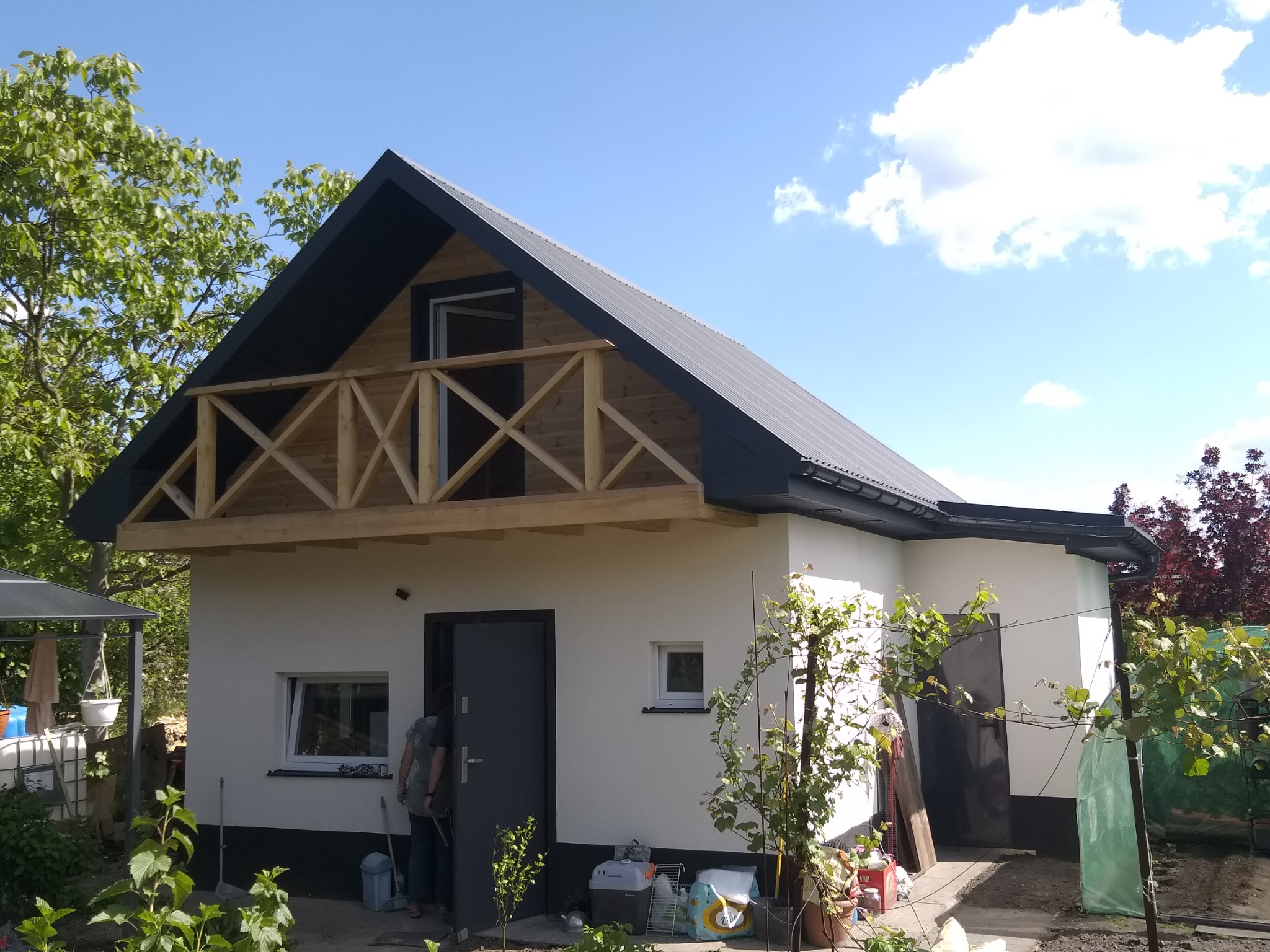 Nowoczesny dom murowany z ciemnym dachem i drewnianym balkonem. Biała elewacja, ciemne rynny. Osoba wchodząca do budynku. Widok na błękitne niebo z chmurami.