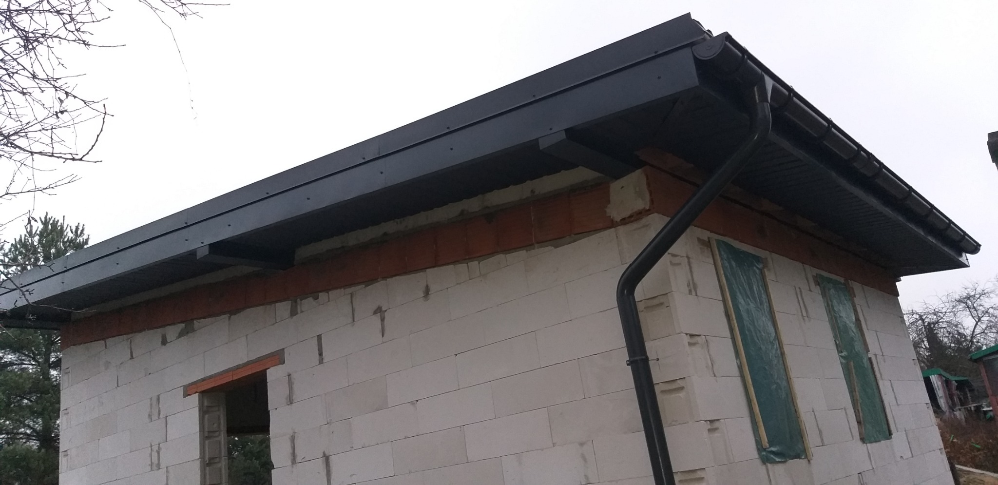 Niski kąt na dom w trakcie budowy z czarnym dachem i rynnami. Okna zabezpieczone folią. Widoczne białe bloczki i brązowe belki.