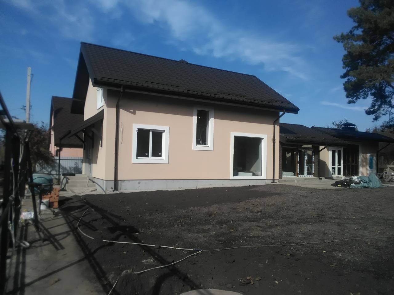 Nowoczesny dom z ciemnym dachem i jasną elewacją w słoneczny dzień. Widoczne rynny, okna z białymi ramami oraz przygotowany teren wokół budynku.