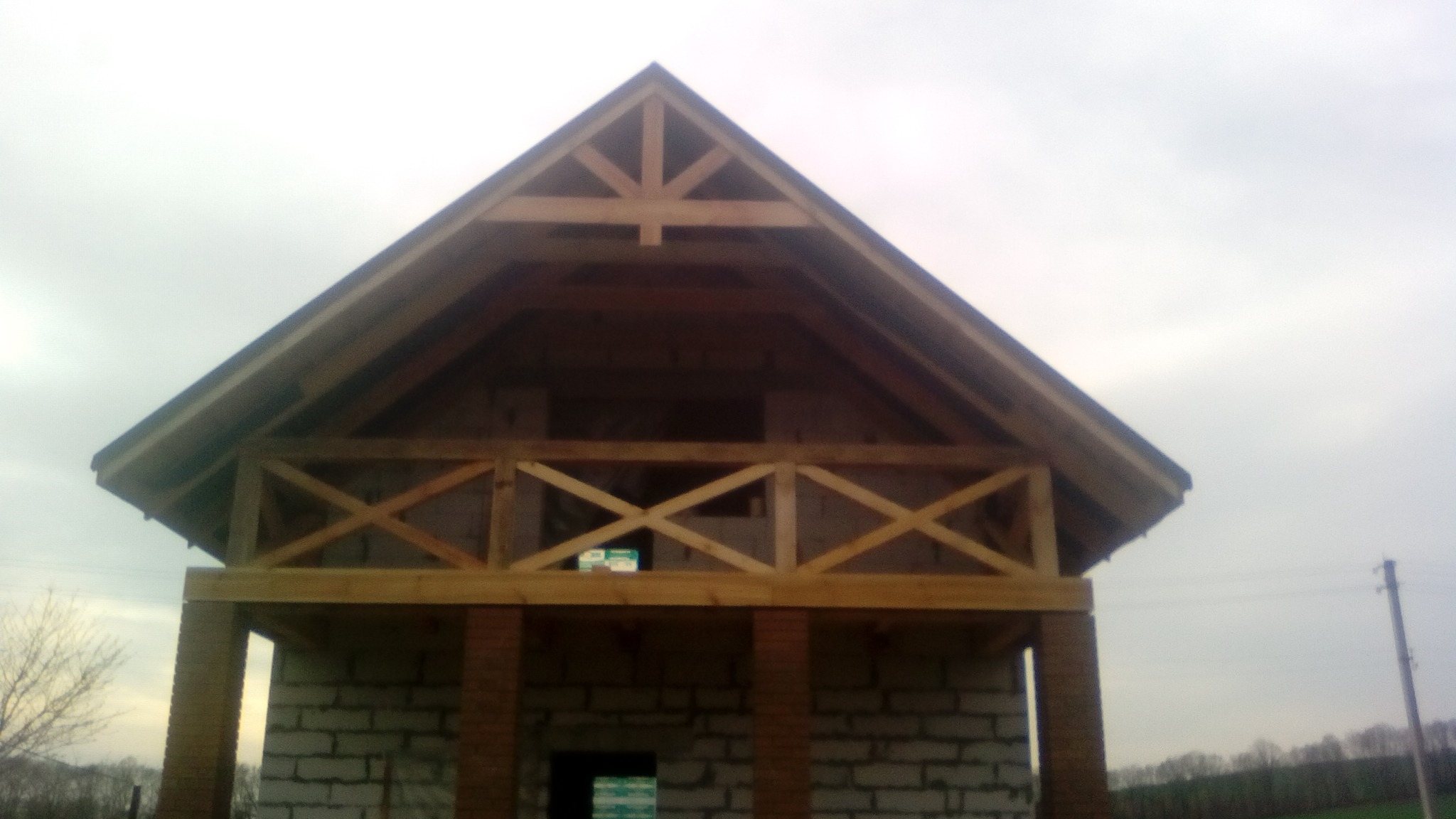 Surowa konstrukcja domu z drewnianą więźbą dachową i balustradą balkonu, wznoszona z bloczków. Widoczne filary z cegły. Tło: pochmurne niebo.