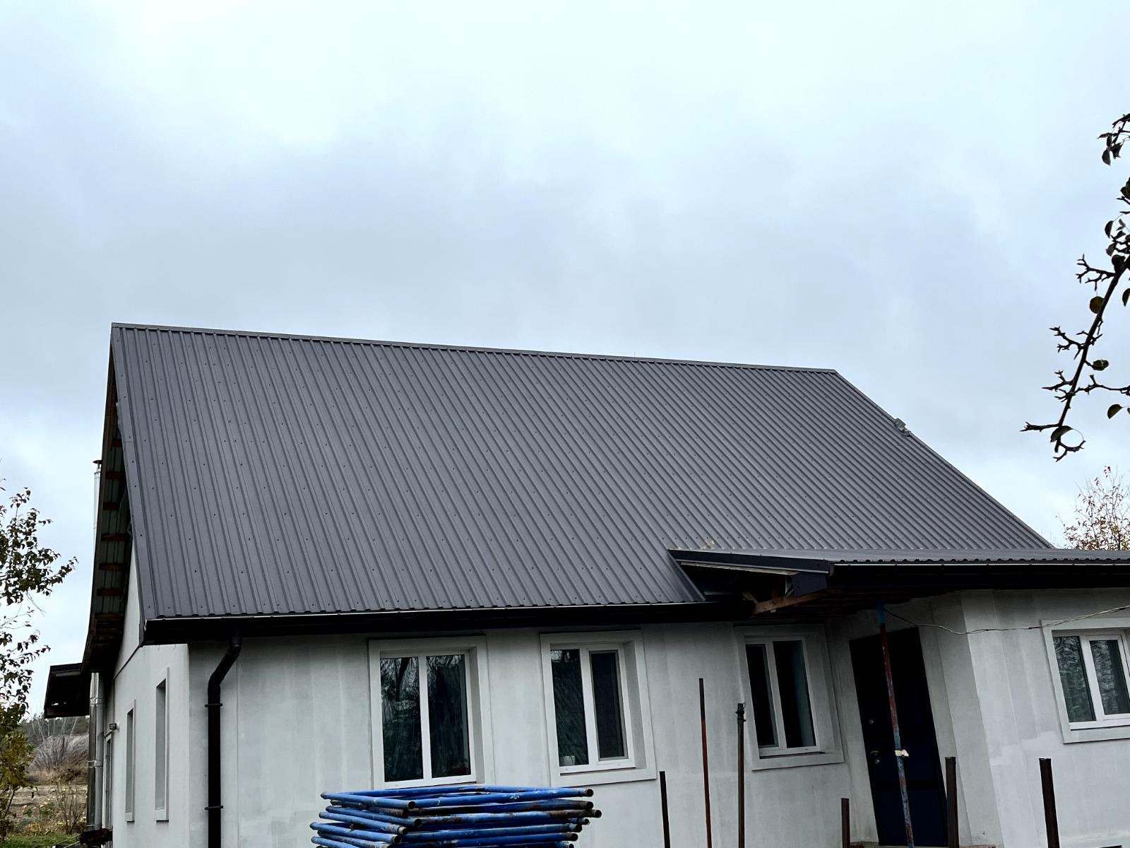 Szary dach z blachy trapezowej na nowym domu z białymi ścianami. Widoczne rusztowania i okna. Pochmurne niebo w tle.